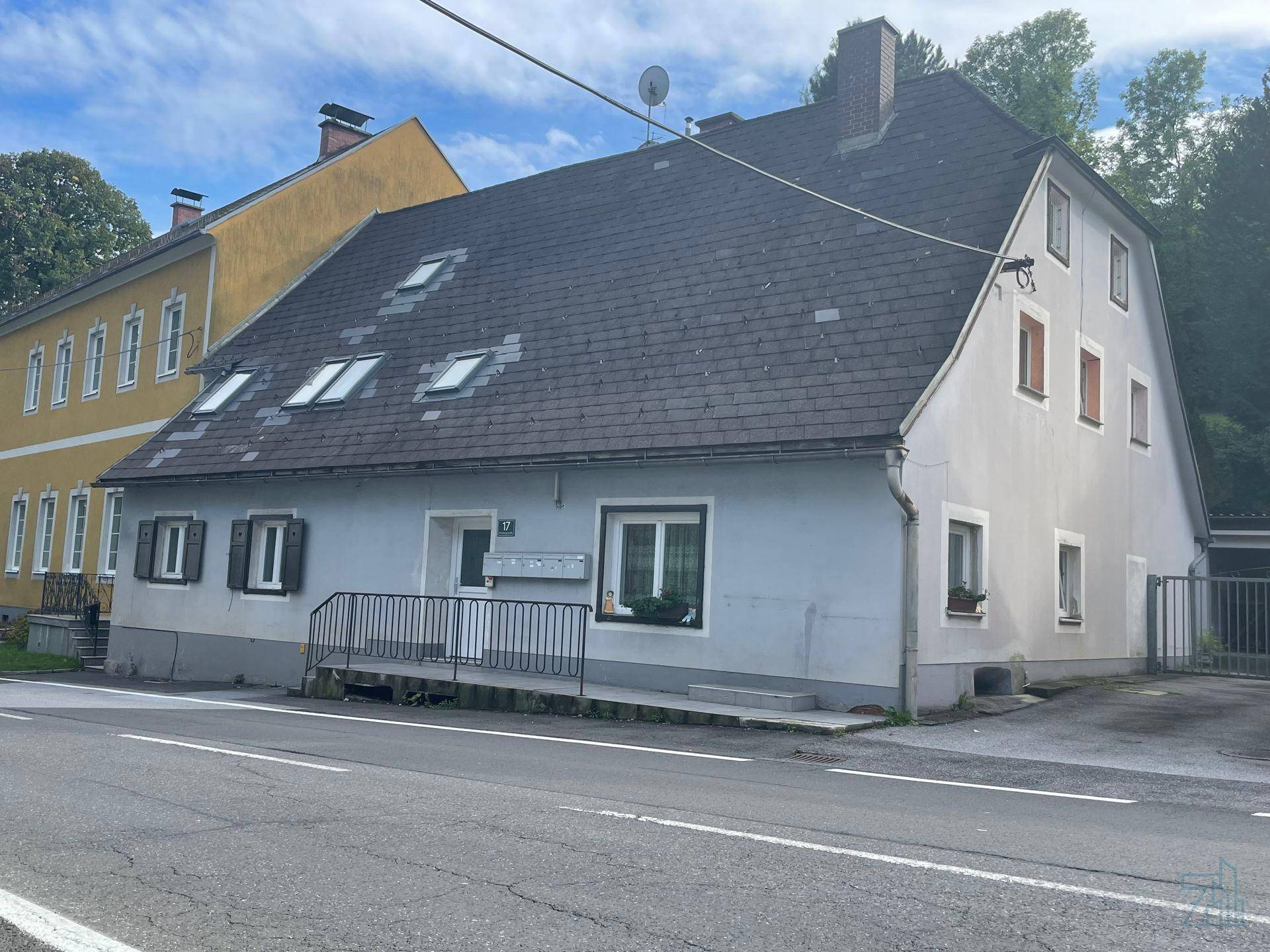 Älteres Wohnhaus mit grauer Fassade, dunklem Dach und mehreren Dachfenstern an einer Straße gelegen.