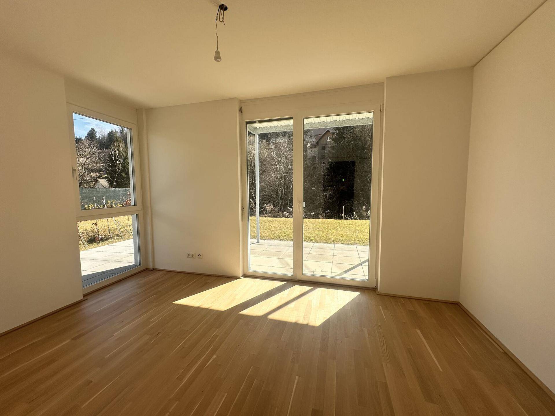 Heller Wohnraum mit Holzparkettboden und großen Fensterfronten, die viel Sonnenlicht hereinlassen und den Blick ins Grüne freigeben.