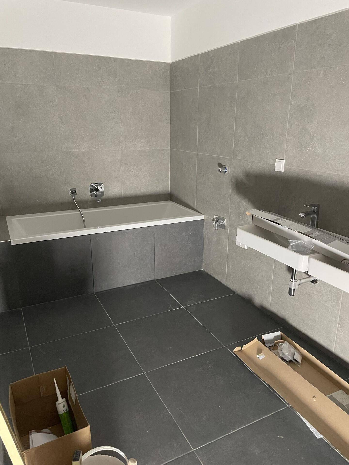 Modernes Badezimmer im Bau mit grauen Fliesen, eingebauter Badewanne und Waschbeckenbereich.