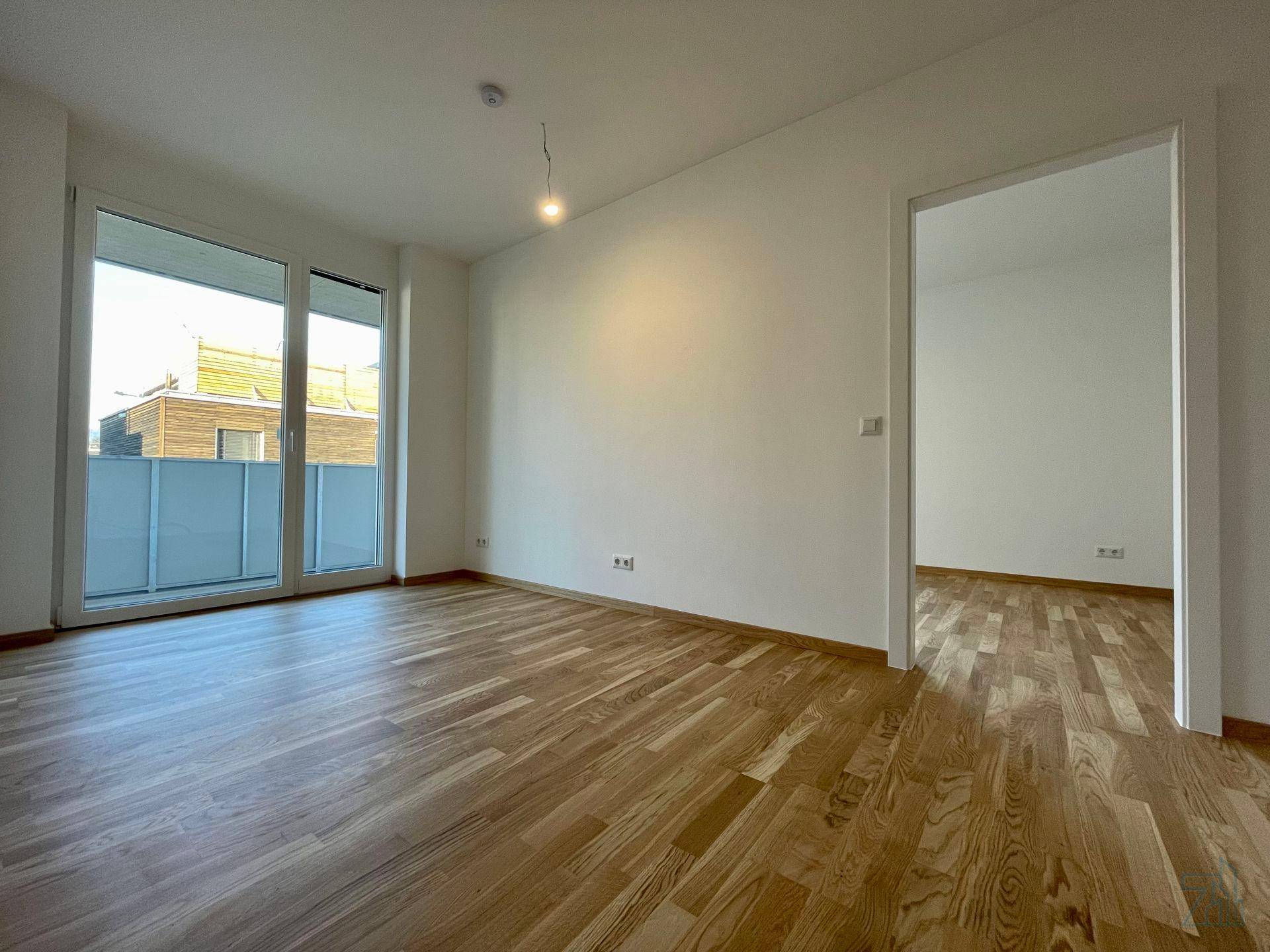 Lichtdurchfluteter Wohnraum mit Holzboden und breiter Balkontür, die viel Tageslicht hereinlässt.