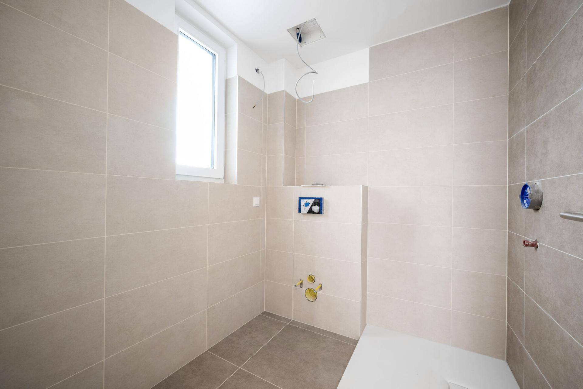 Unfertiges Badezimmer mit beigefarbenen Fliesen, Fenster und vorbereiteten Anschlüssen.