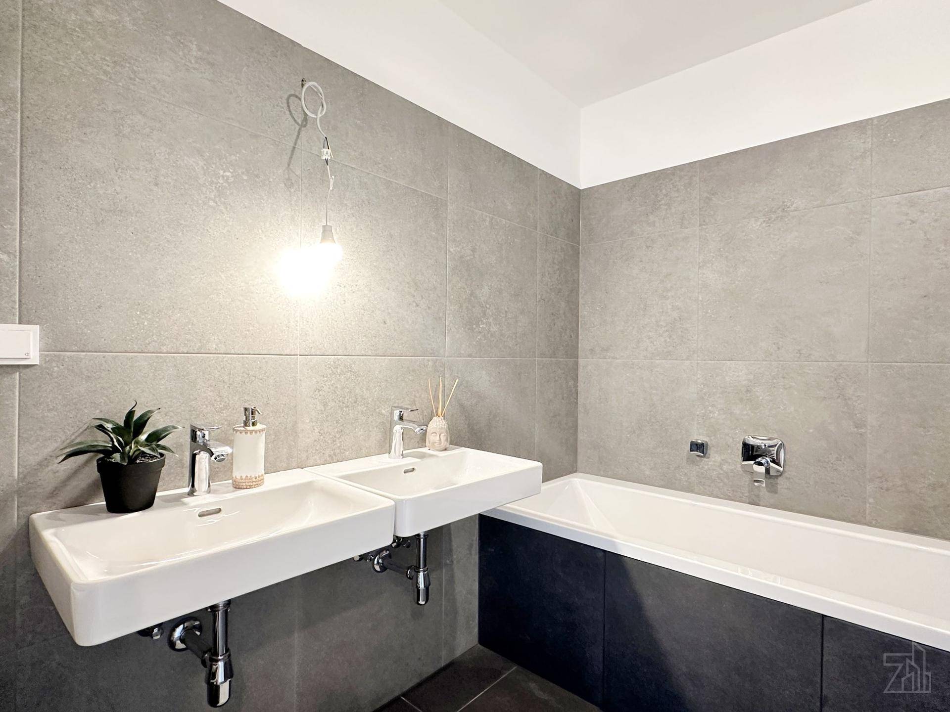 Modernes Badezimmer mit Doppelwaschbecken, Badewanne und großformatigen grauen Fliesen.