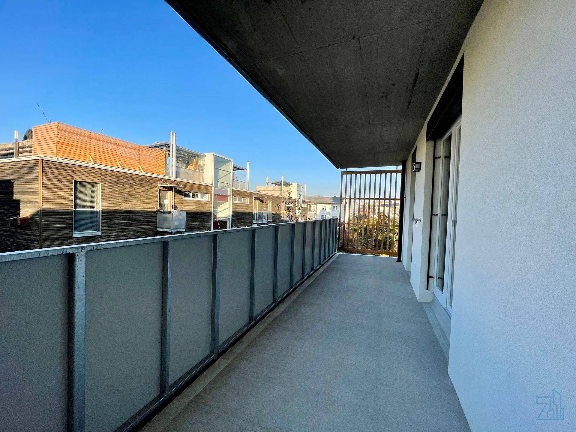 Langer Balkon mit Glasbrüstung und Blick auf die umliegenden Gebäude unter blauem Himmel.