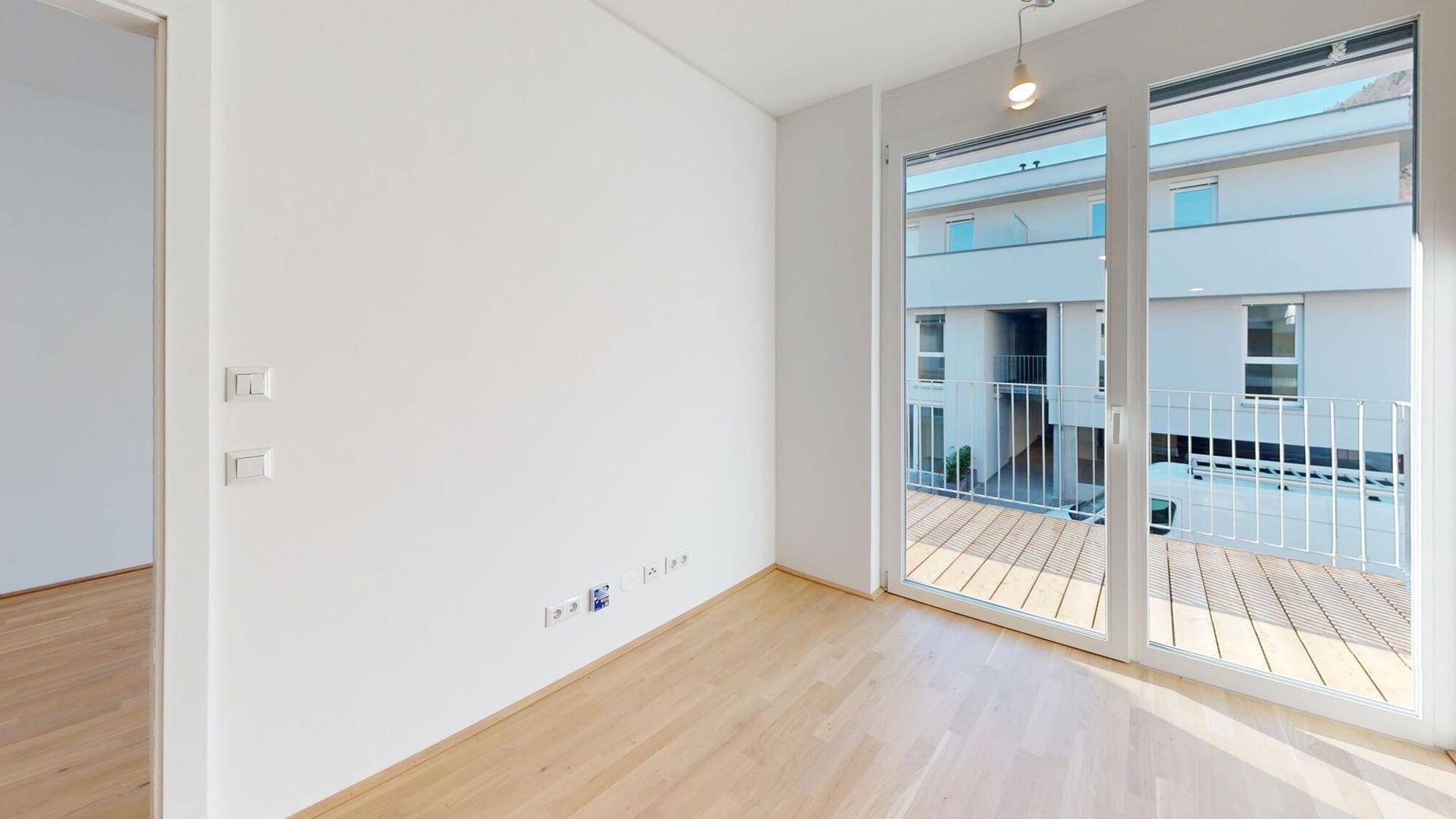 Lichtdurchfluteter Raum mit Holzboden, weißen Wänden und direktem Zugang zu einem Balkon.