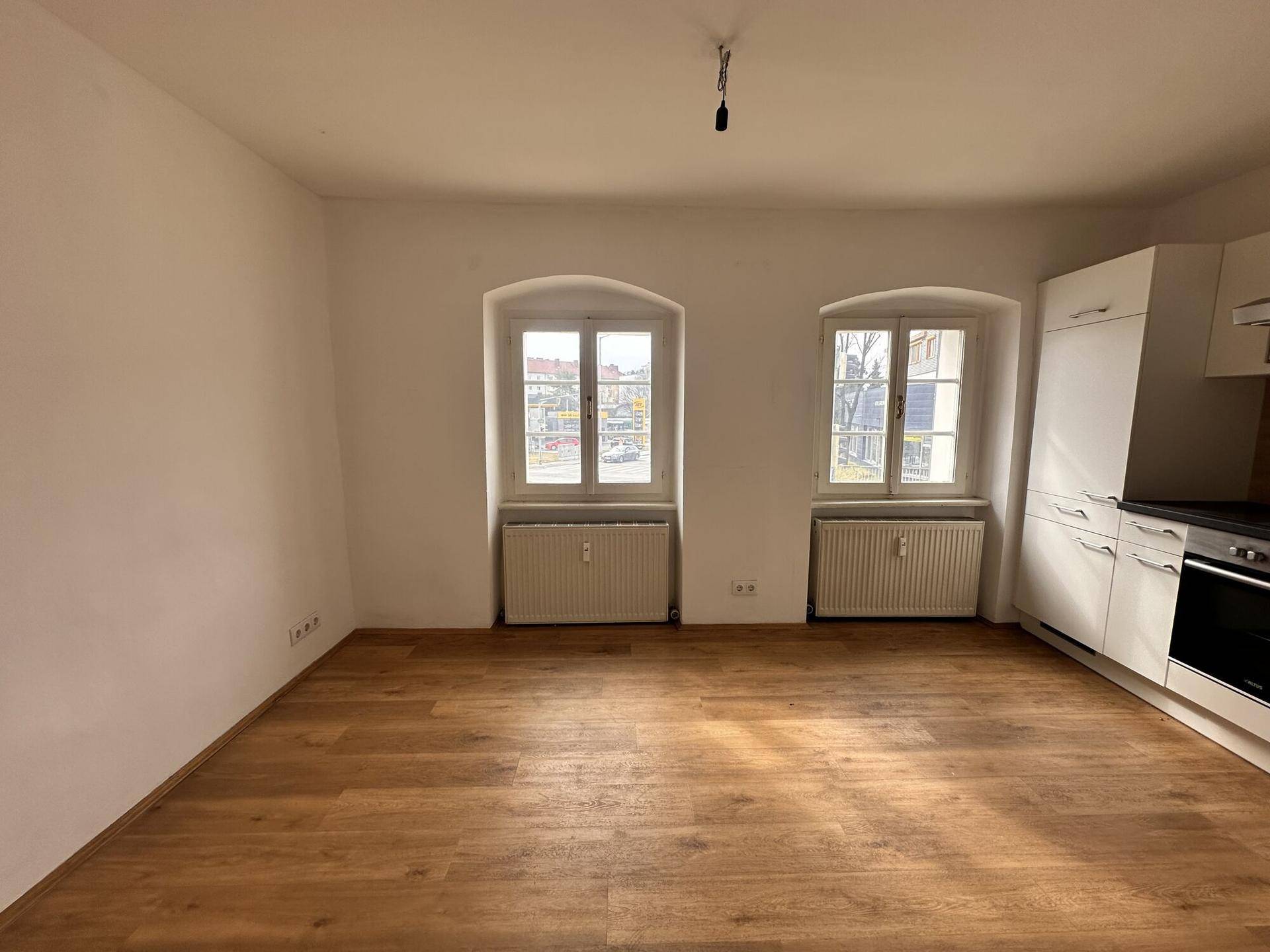 Offener Wohnbereich mit integrierter Küchenzeile, Holzboden und zwei Fenstern.