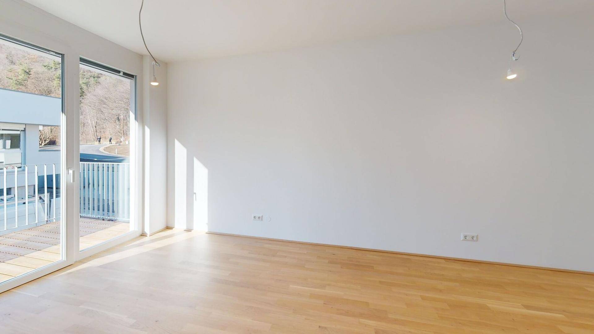 Heller, leerer Wohnraum mit großen Fenstern, Holzboden und Zugang zu einem sonnigen Balkon.