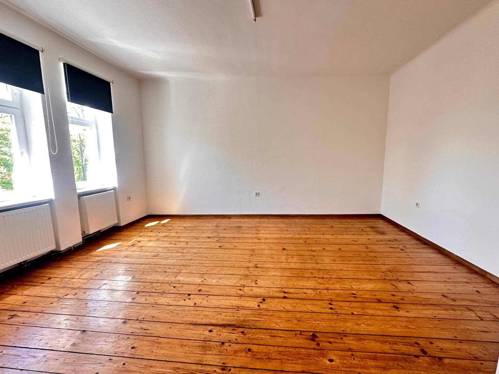 Leerer Raum mit Holzdielenboden, weißen Wänden und zwei Fenstern mit Heizkörpern.