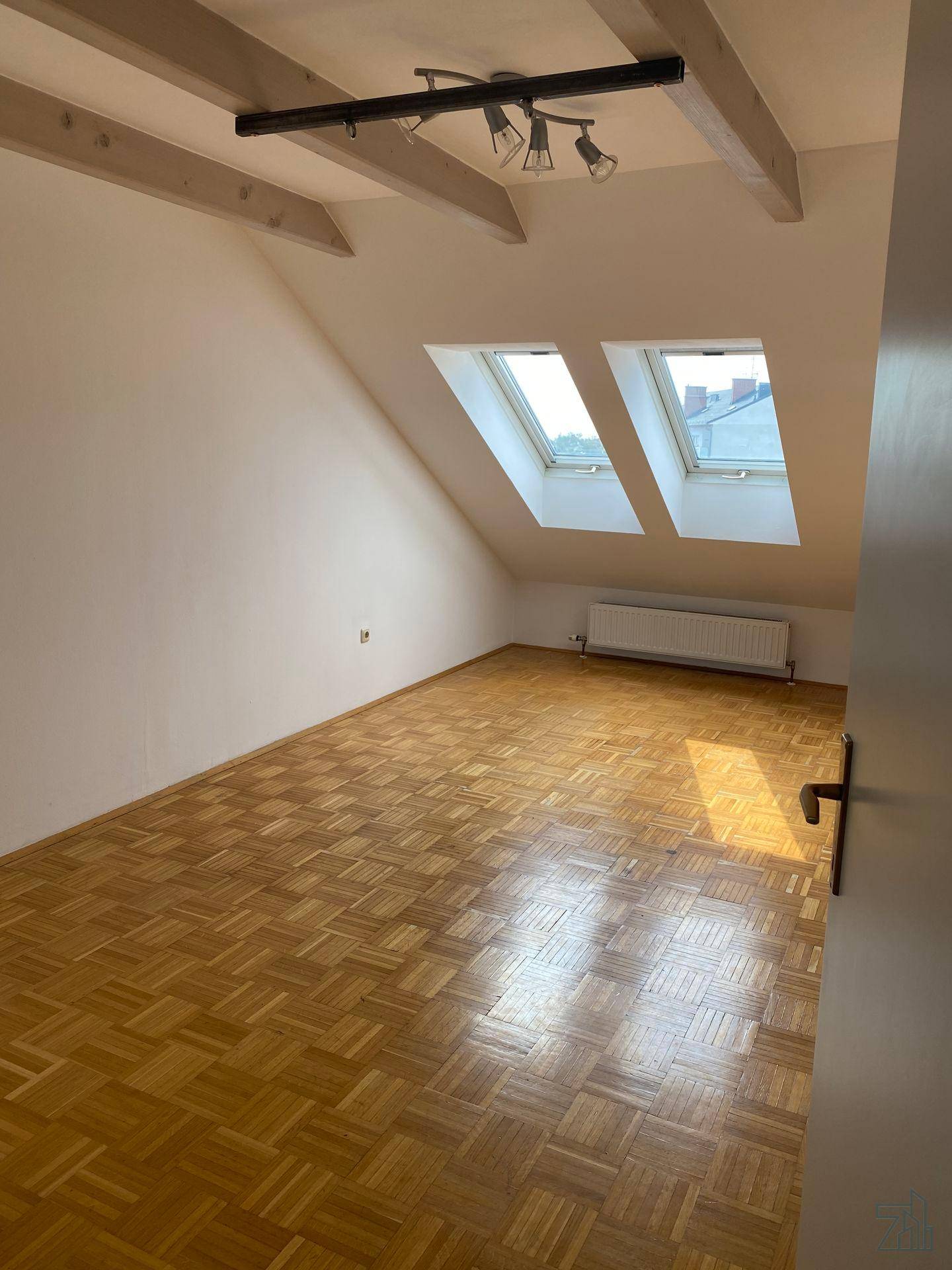 Helles Dachzimmer mit Holzbalken, Parkettboden und zwei Dachfenstern, ideal als Schlaf- oder Wohnbereich.
