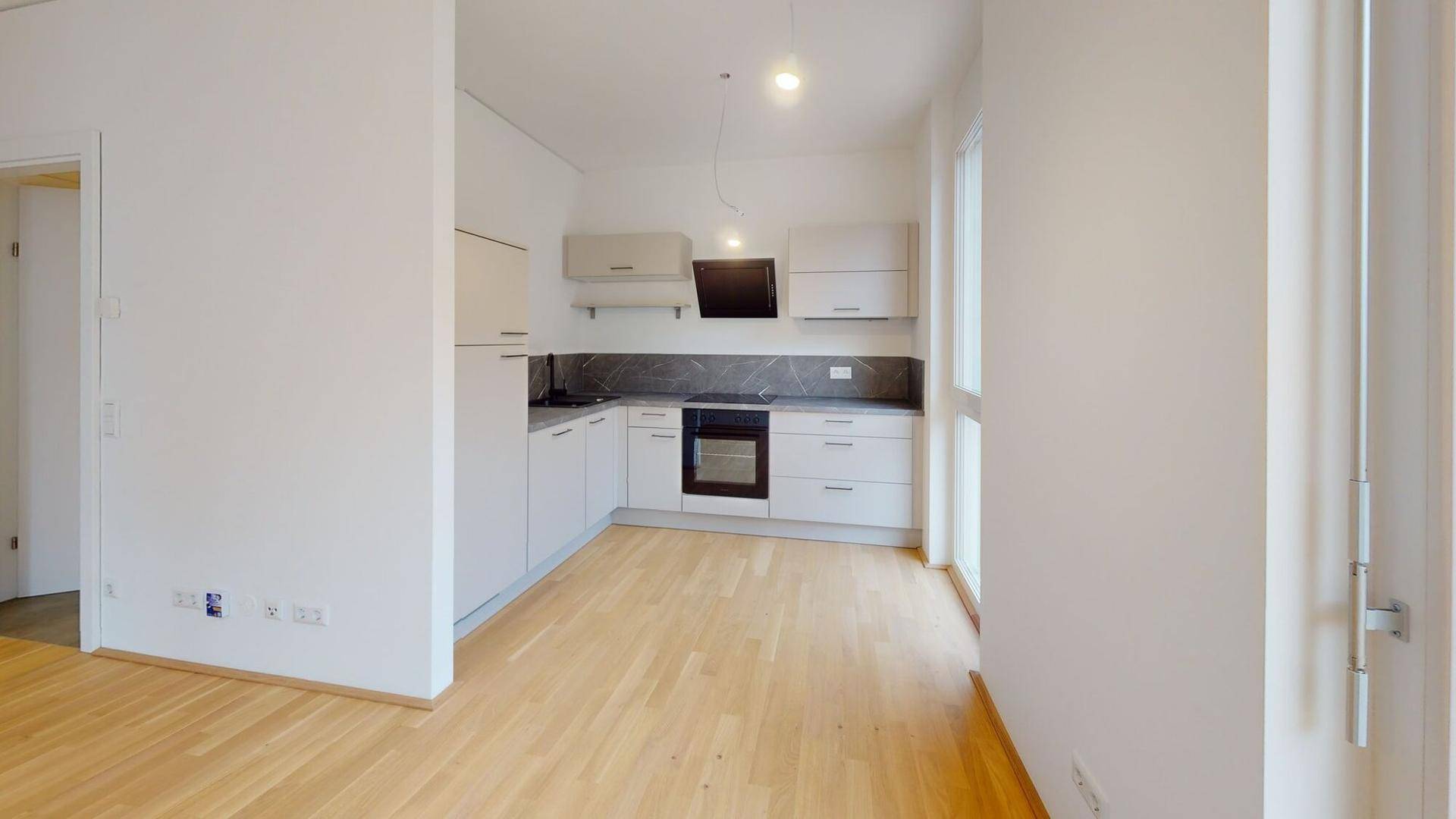 Helle, leere Küche mit modernen weißen Schränken, Holzboden und Zugang zum Balkon.