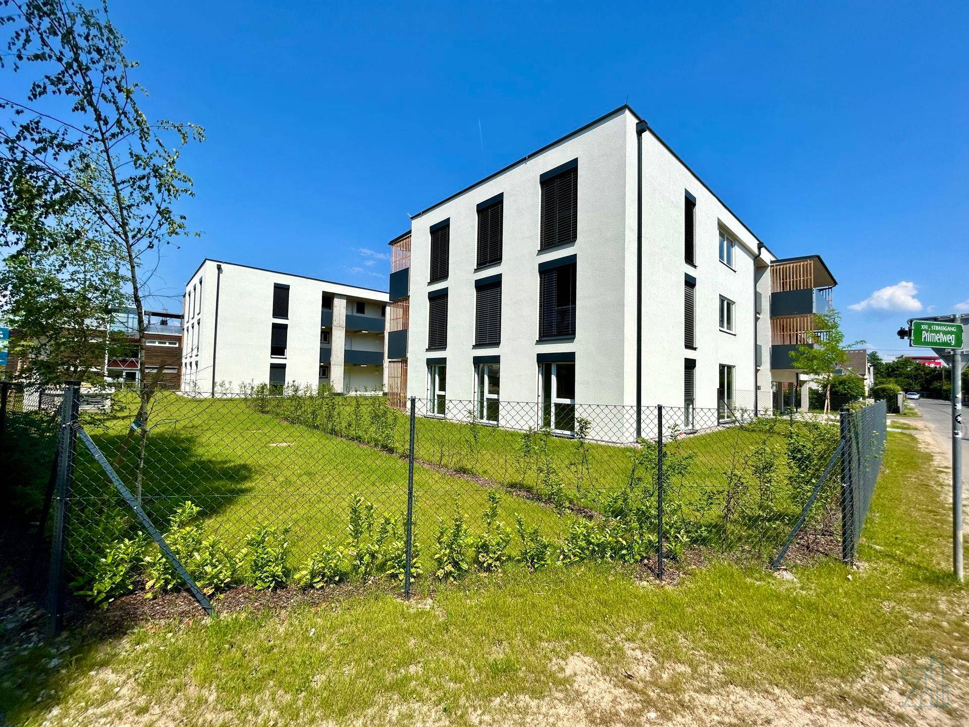 Zwei moderne Wohngebäude mit klarer Architektur, umgeben von Grünflächen und einem Zaun.