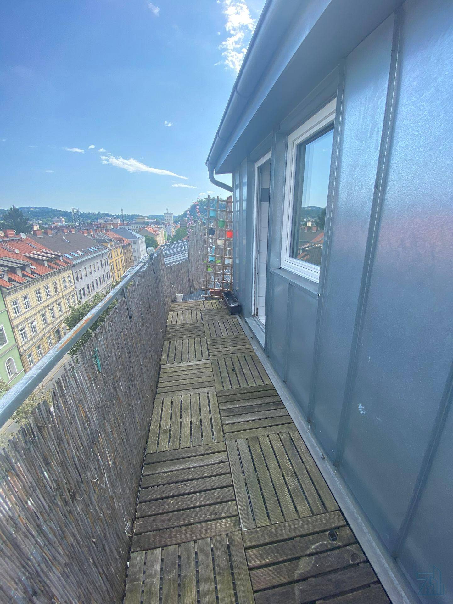 Schmaler Balkon mit Holzboden und Sichtschutz, bietet einen weiten Stadtblick unter blauem Himmel.