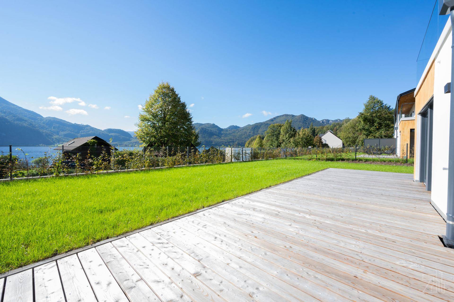 Geräumige Holzterrasse grenzt an einen grünen Rasen mit Panoramablick auf den See und die umliegenden Berge.