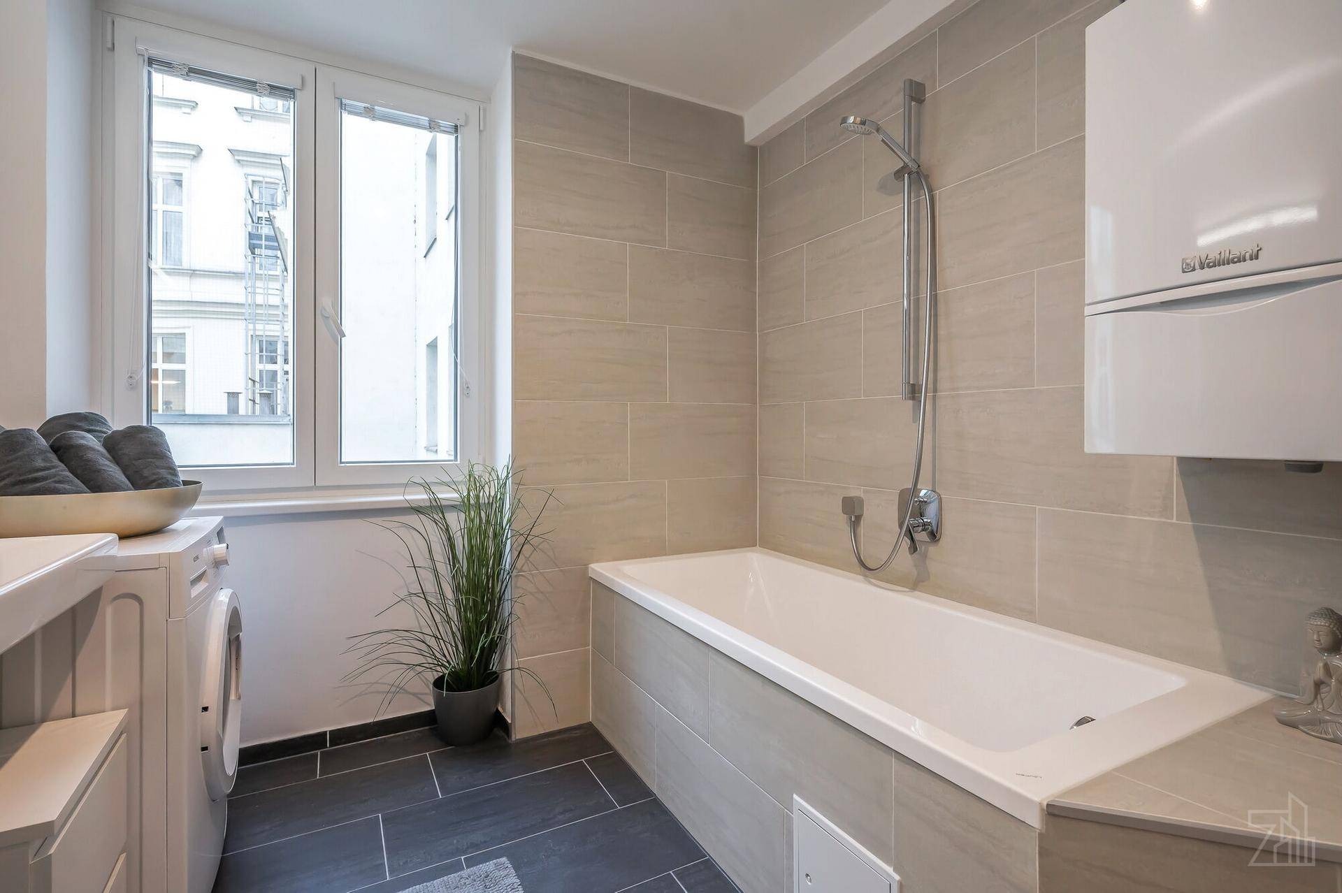 Helles Badezimmer mit Badewanne, Fenster und Waschmaschine, ausgestattet mit modernen Fliesen.