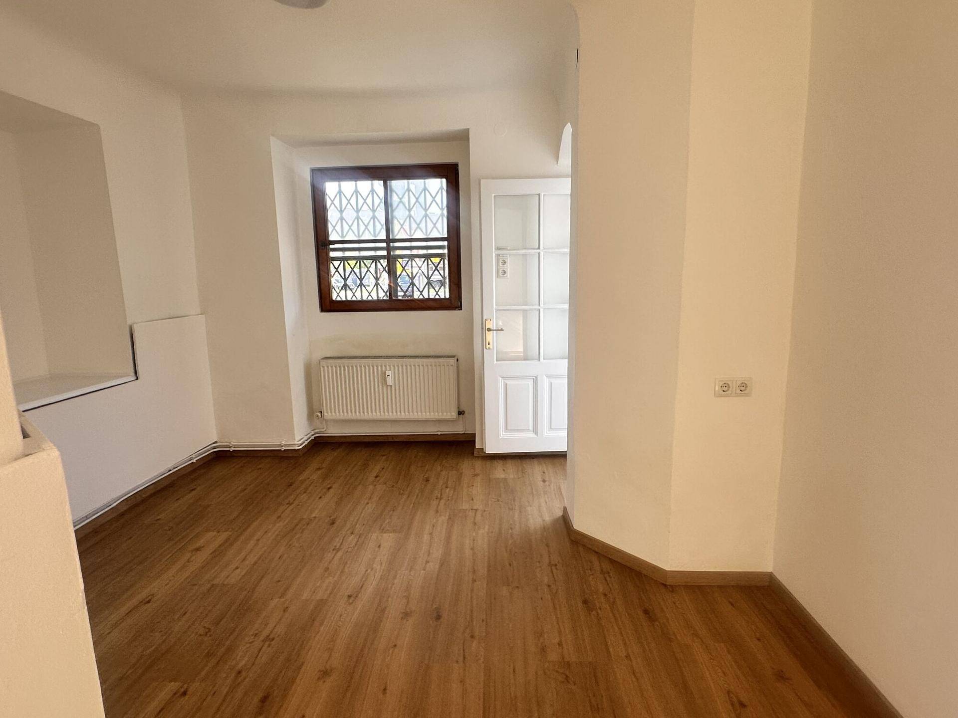 Heller Raum mit Holzboden, einem dekorativen Fenster und Zugang zu einem weiteren Bereich.