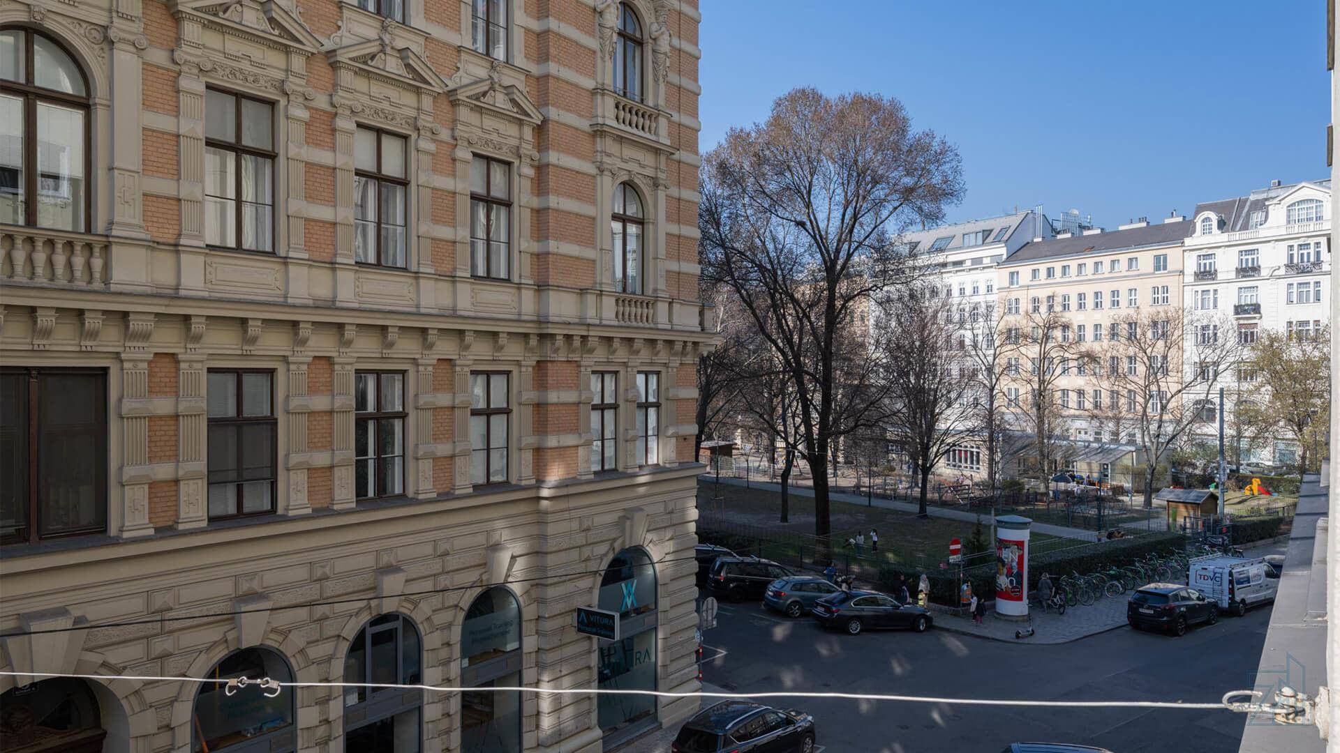 Historische Gebäude entlang einer Stadtstraße mit geparkten Autos und Bäumen im Vordergrund.