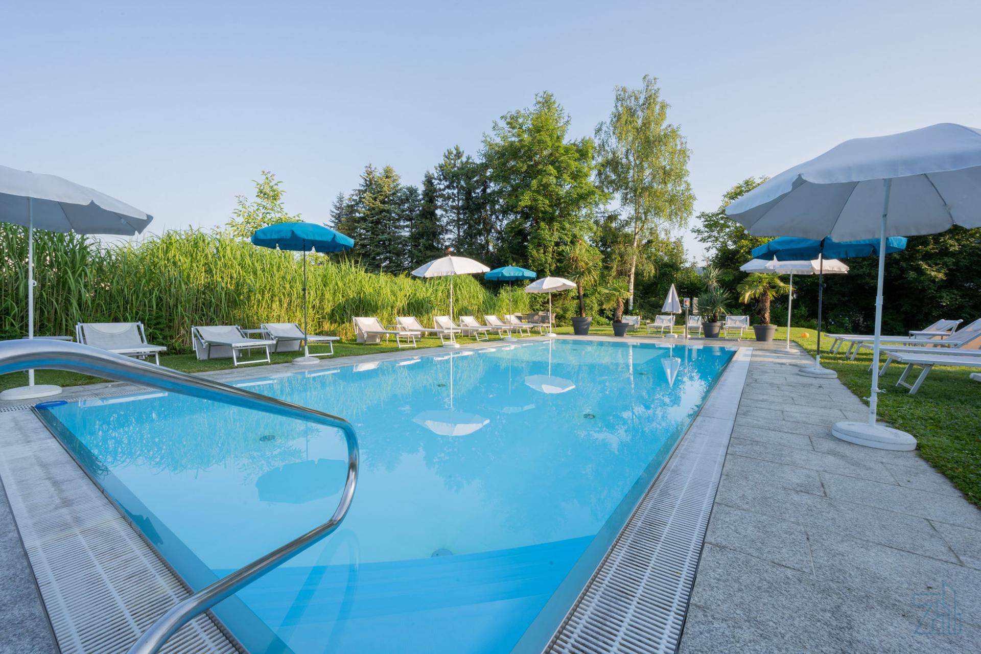 Weitläufiger Poolbereich mit vielen Liegestühlen und Sonnenschirmen, umgeben von Grün.