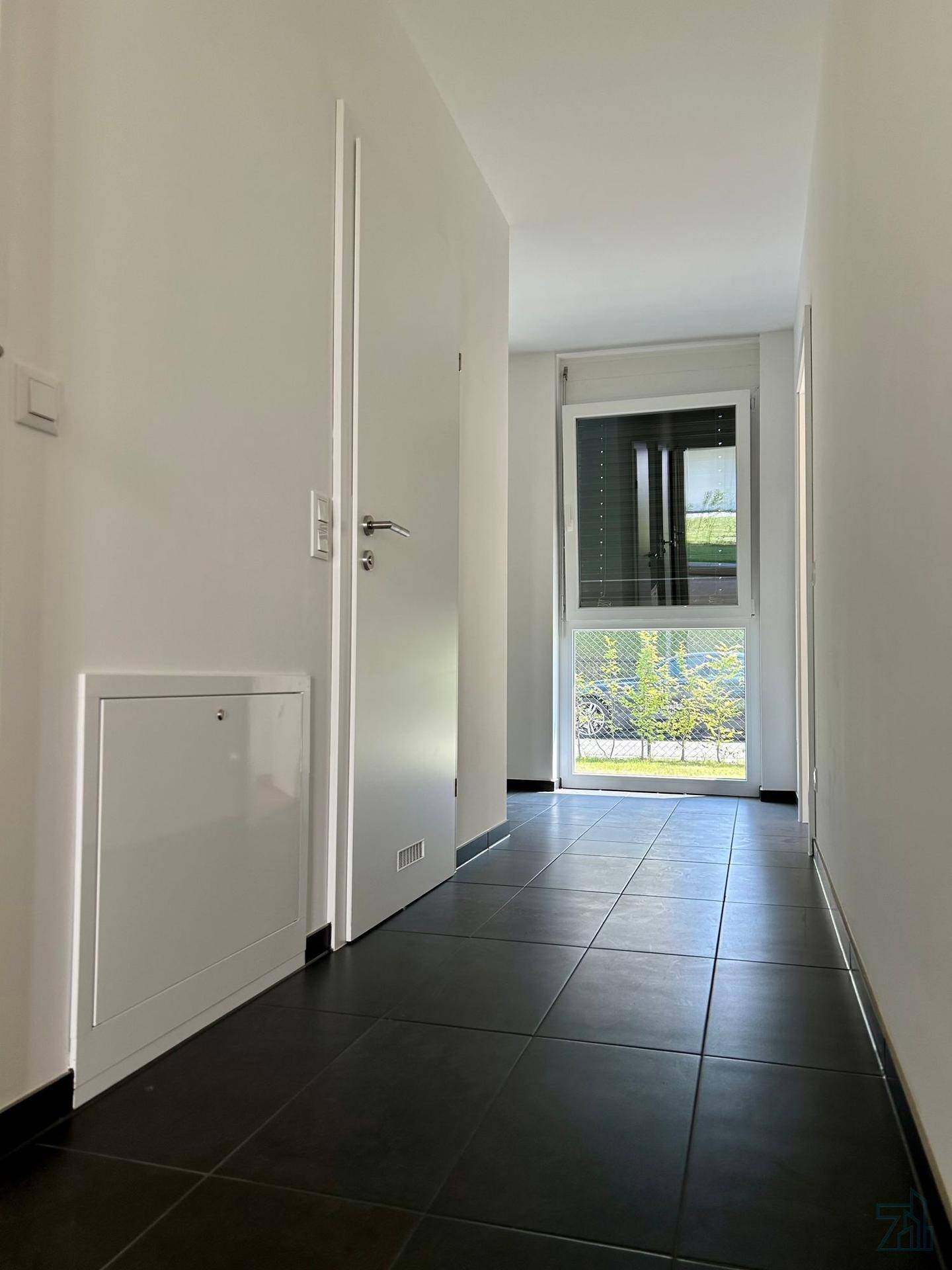 Moderner Flur mit dunklen Fliesen und einem Fenster am Ende, das Tageslicht spendet.