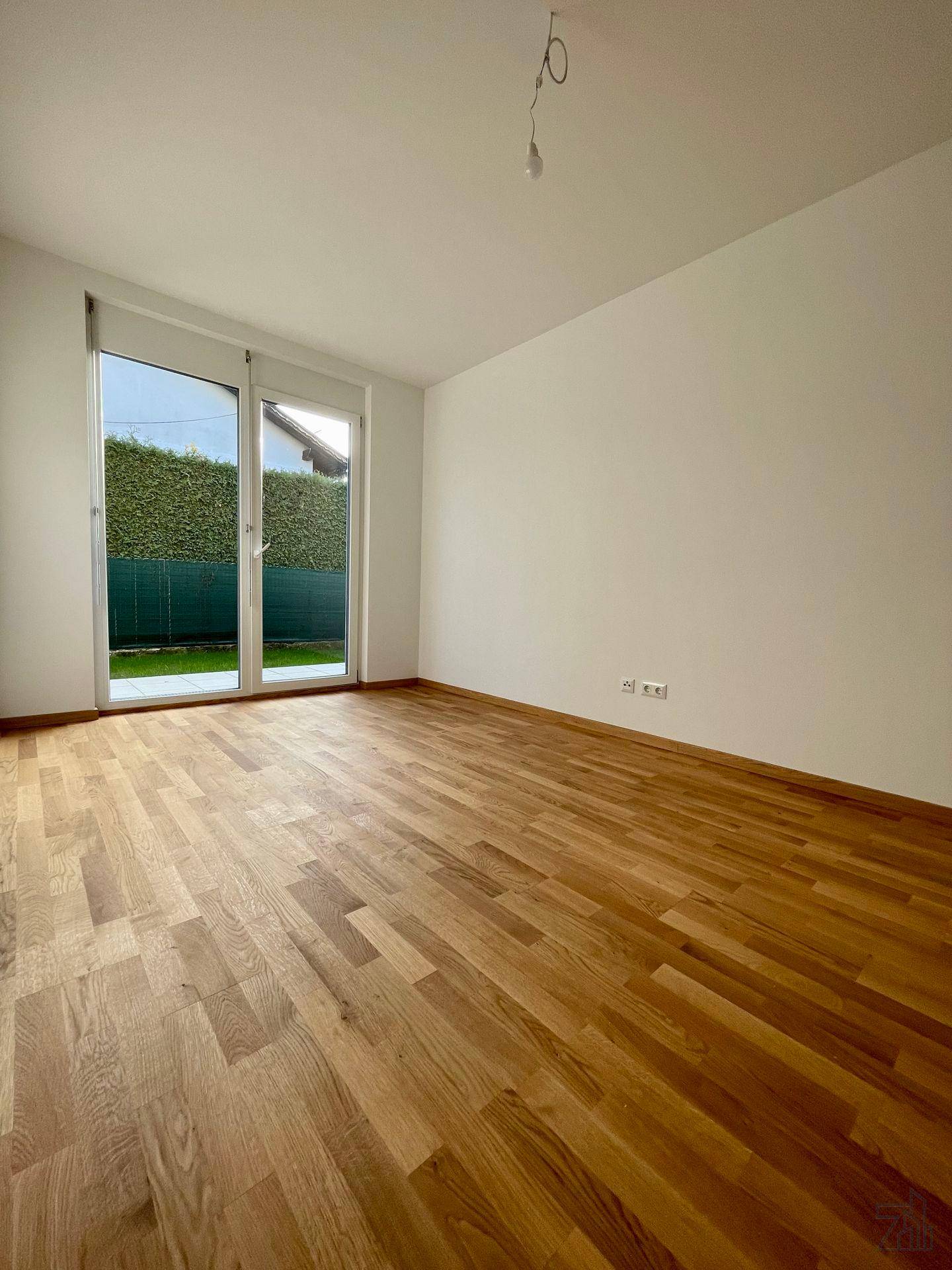 Helles, leeres Zimmer mit Holzboden und großer Glastür, die zu einem grünen Gartenbereich führt.