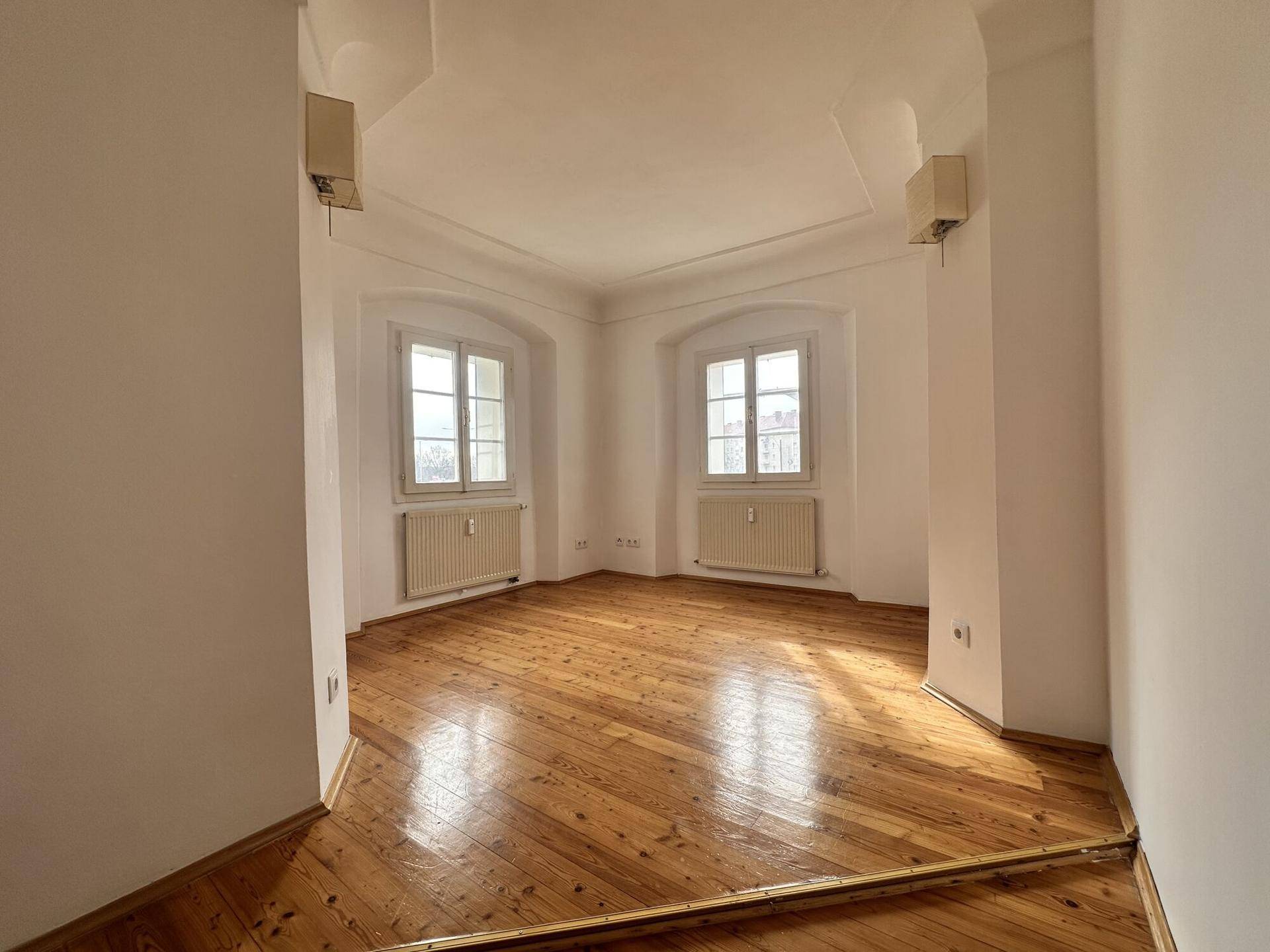 Heller Raum mit Holzboden, zwei Fenstern und einer dekorativen Decke.