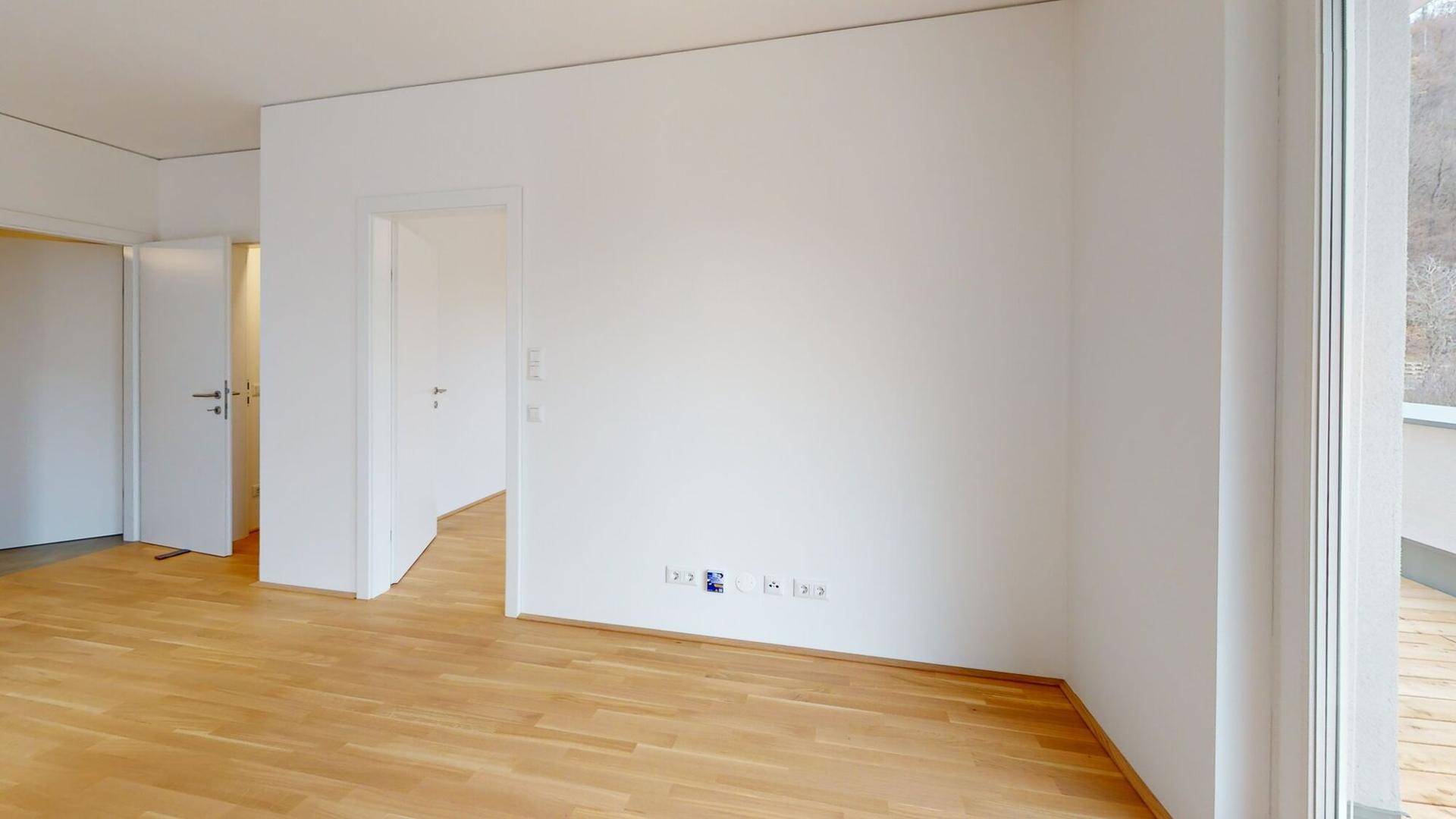 Leeres Zimmer mit hellem Holzboden, weißen Wänden und mehreren Türöffnungen sowie Balkonzugang.