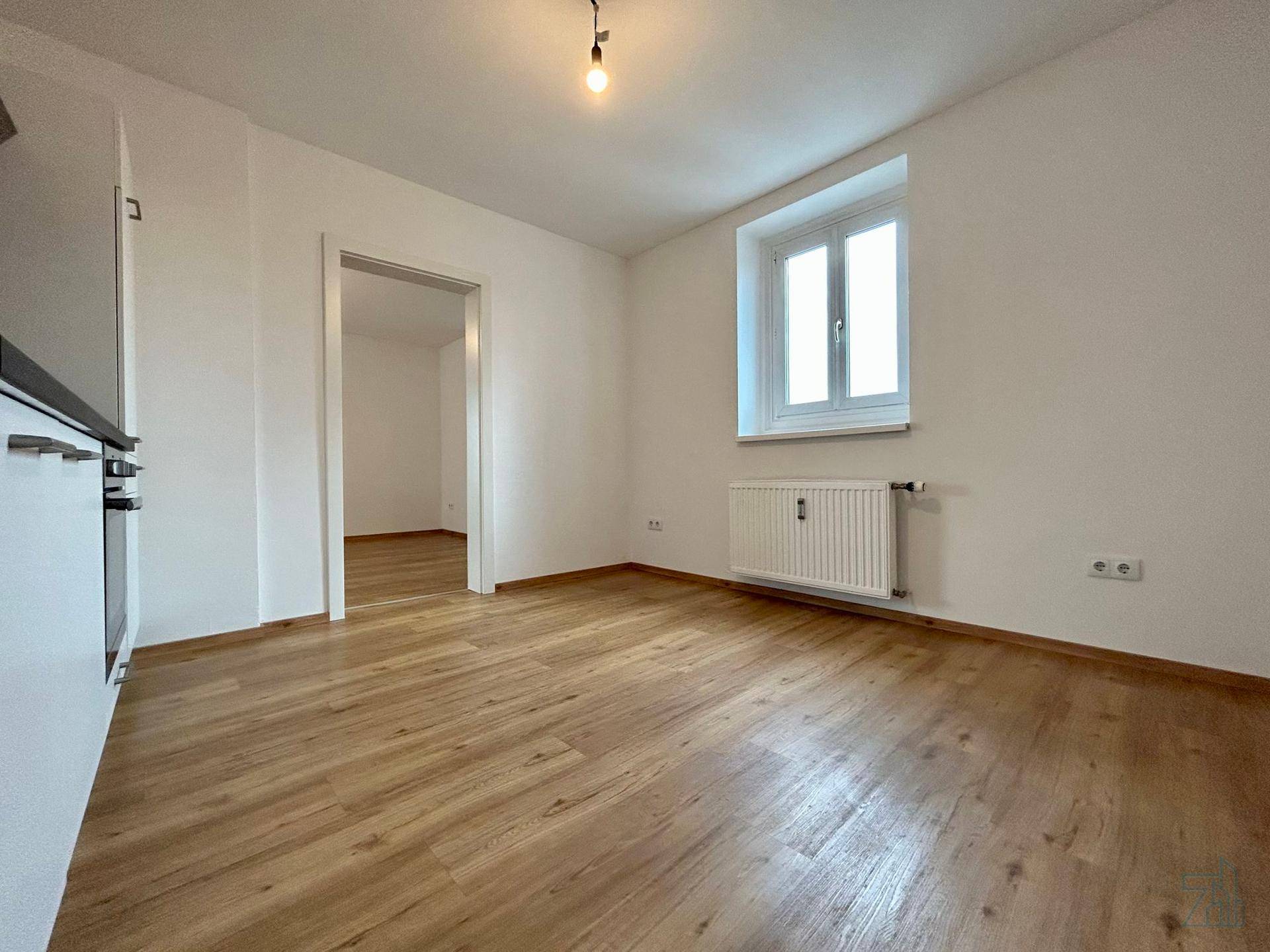 Heller Wohnbereich mit Laminatboden, weiß gestrichenen Wänden und einem Fenster mit Heizkörper.