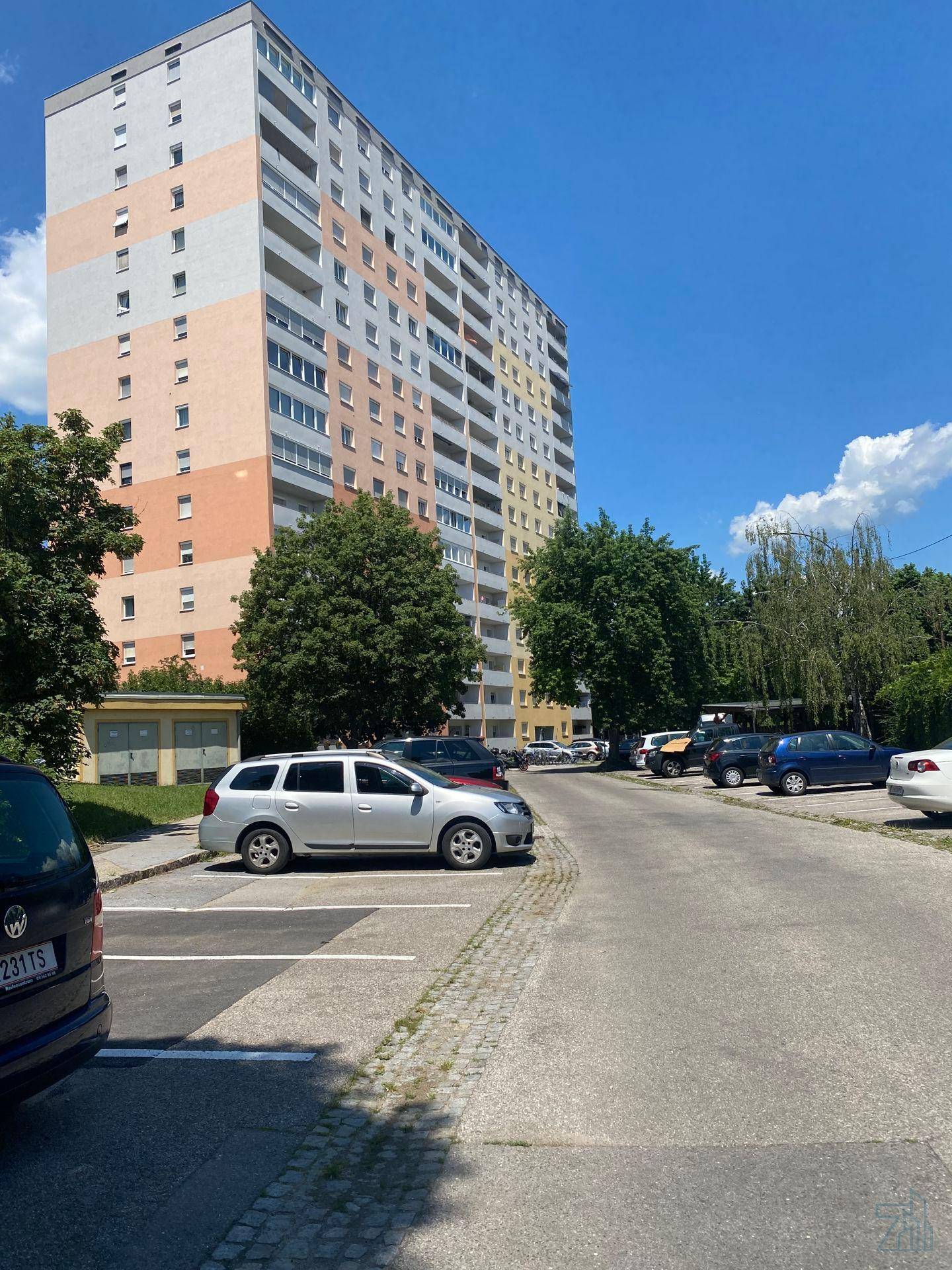 Hohes Wohngebäude mit vielen Etagen und Parkplätzen davor unter blauem Himmel.