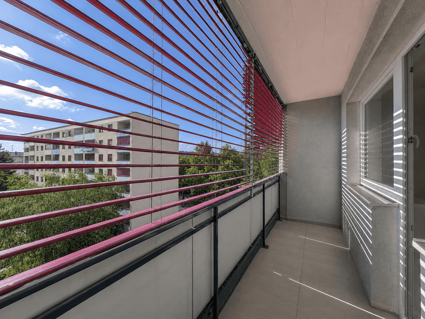 Heller Balkon mit roten Jalousien und Blick auf die umliegenden Gebäude und Bäume.