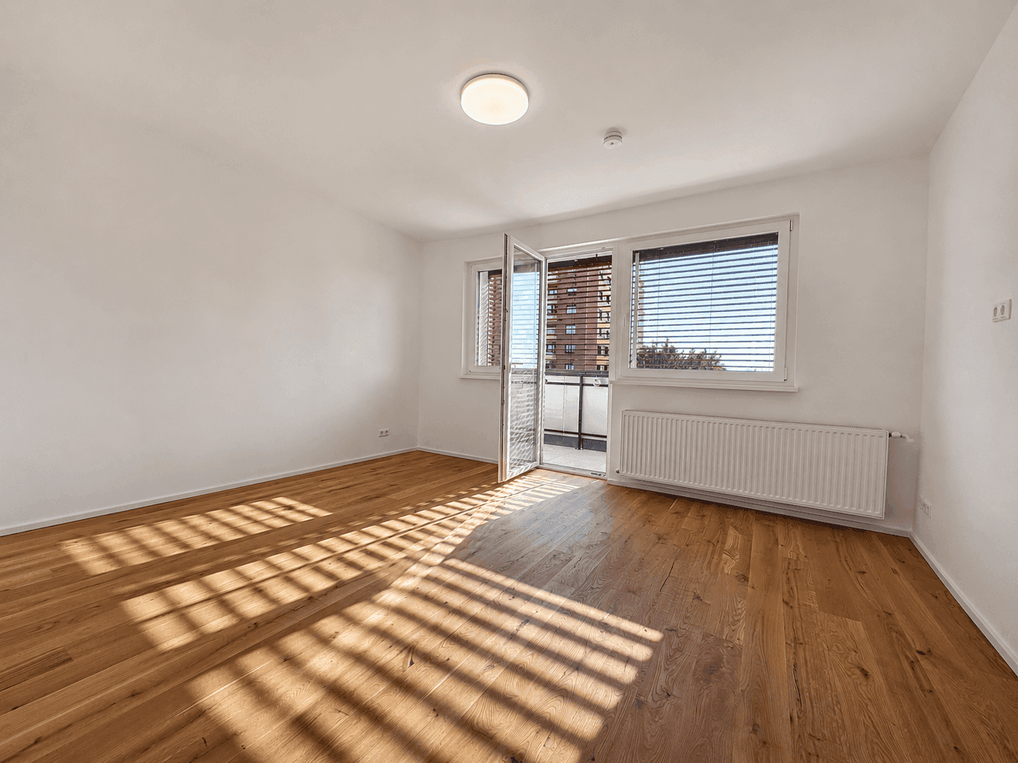 Lichtdurchflutetes Wohnzimmer mit Holzboden und offener Balkontür, die frische Luft hereinlässt.