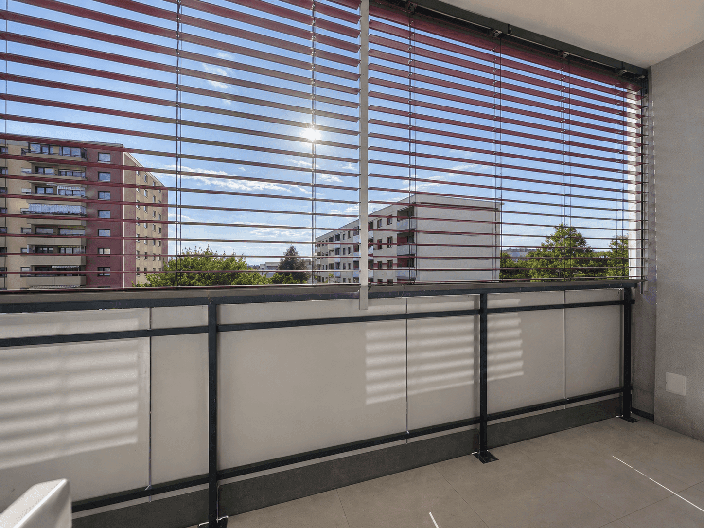 Geräumiger Balkon mit roten Jalousien und sonnigem Ausblick auf die Nachbarschaft.