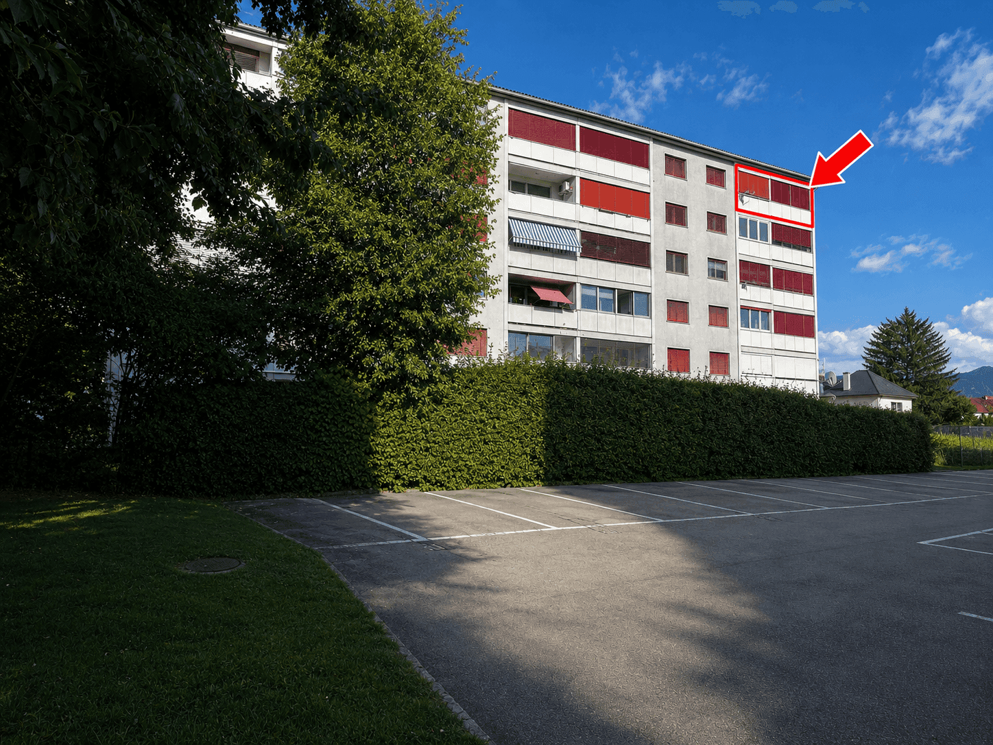 Mehrfamilienhaus mit Balkonen und Parkplatz, umgeben von Bäumen und Hecken.