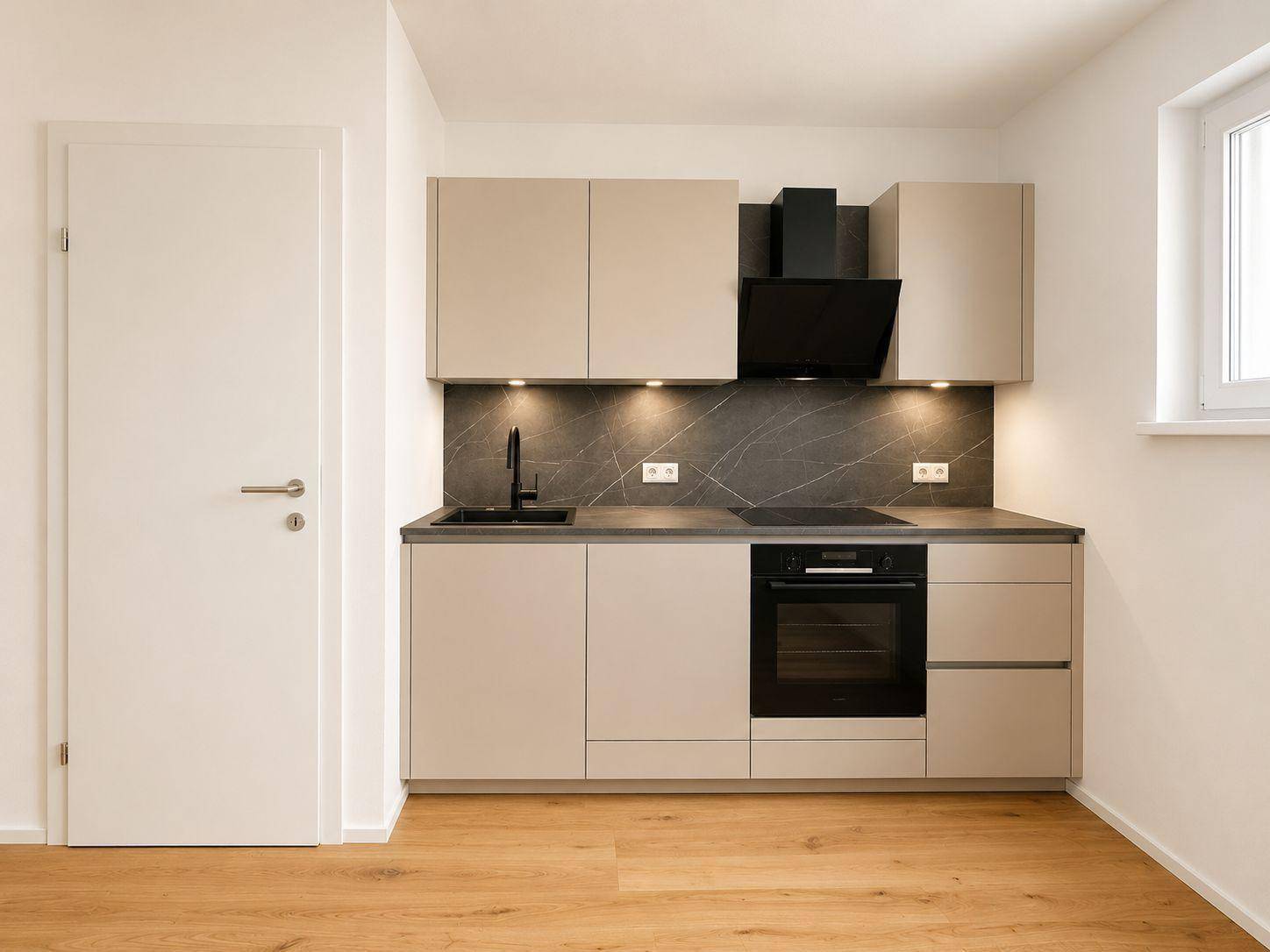 Kompakte, moderne Einbauküche mit hellen Fronten und dunkler Arbeitsplatte, neben einer Tür.