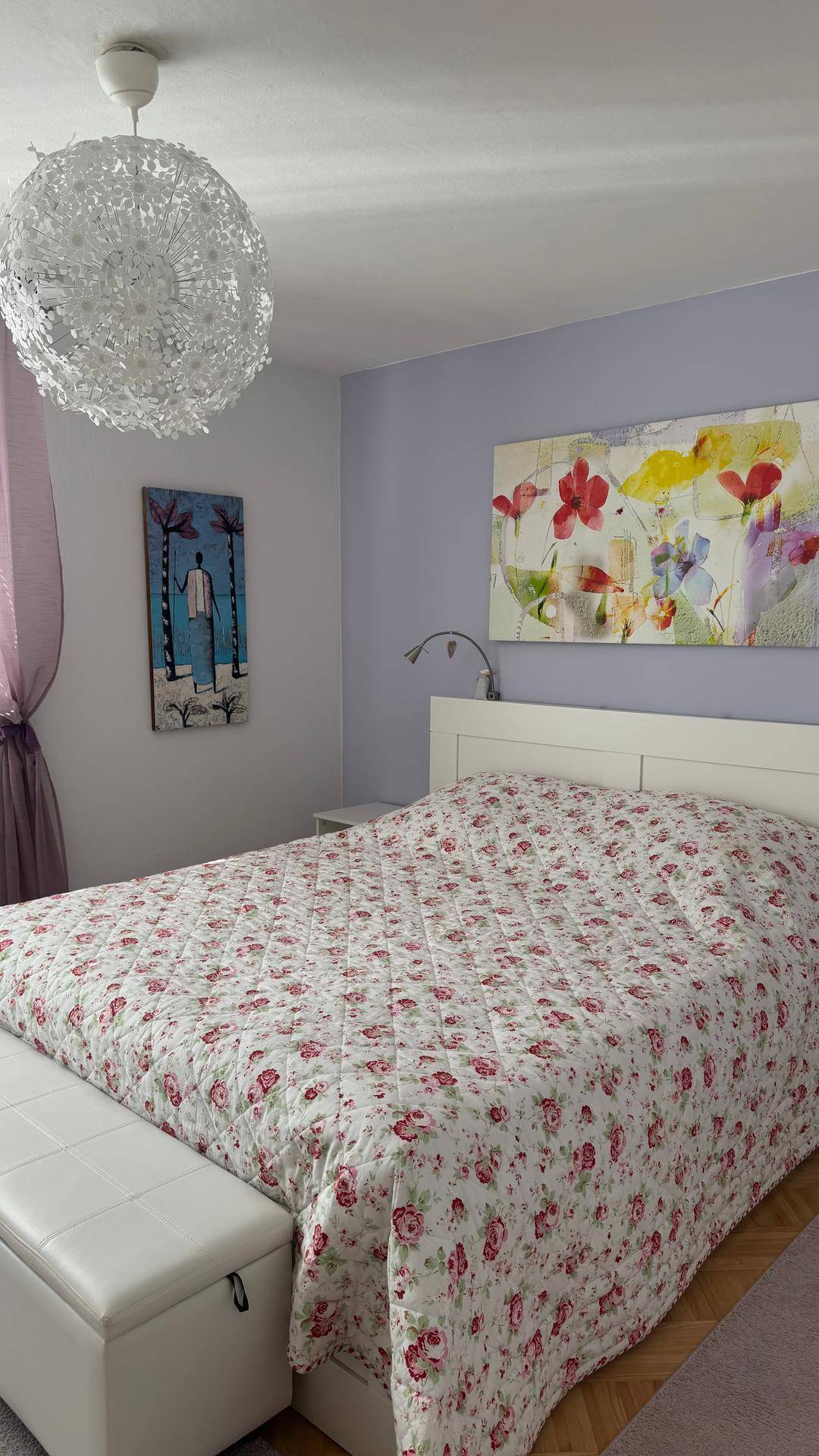 Ruhiges Schlafzimmer mit floraler Bettwäsche, lila Wand und dekorativer Deckenleuchte.
