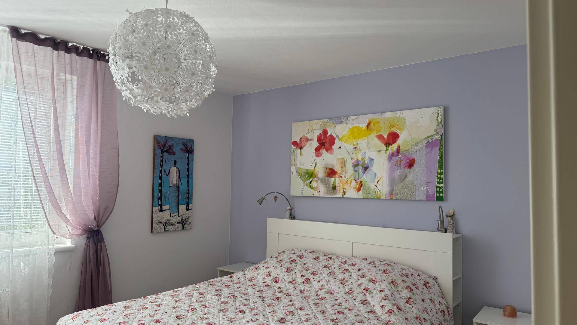 Helles Schlafzimmer mit floraler Bettwäsche, lila Wandakzent und modernem Deckenlicht.
