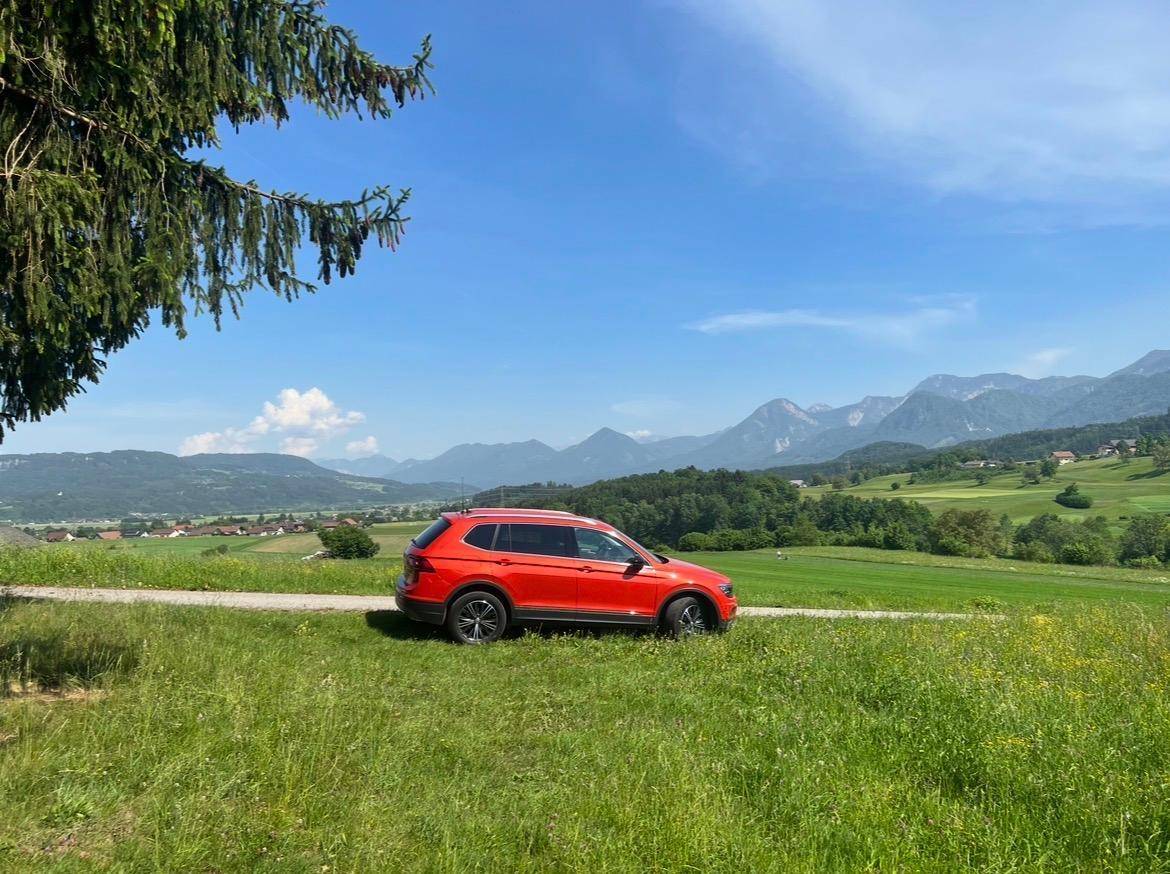 Rotes Auto auf einem Feldweg mit weitem Blick über die grüne Landschaft und die majestätischen Berge.