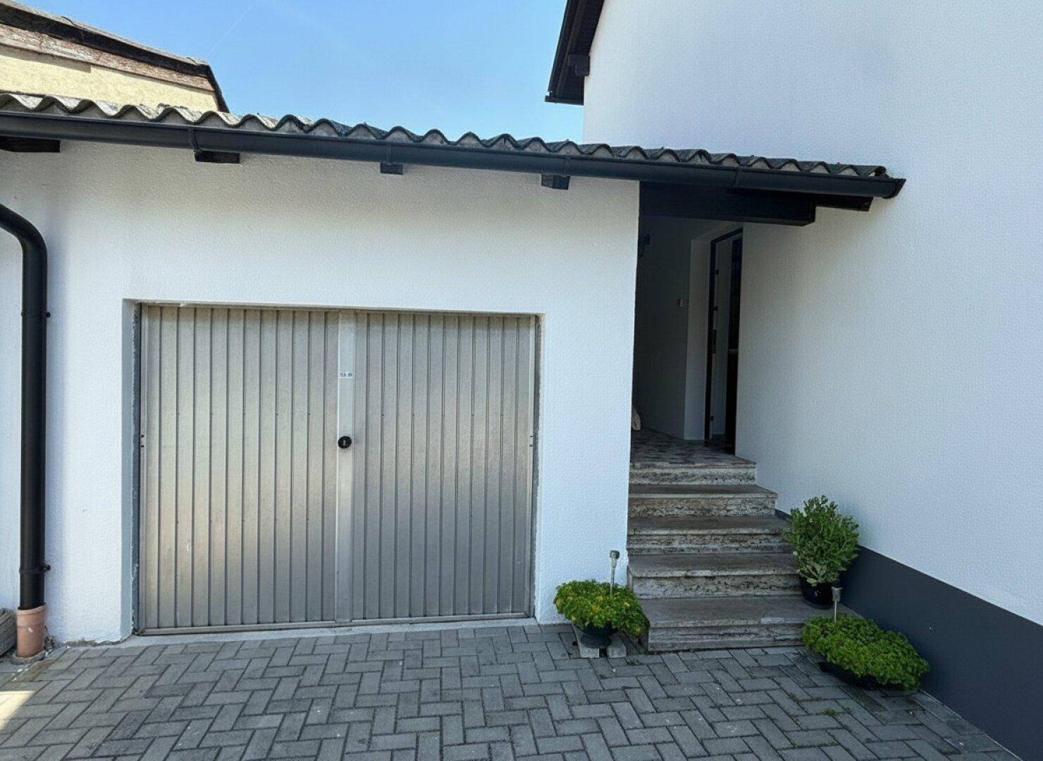 Eingangsbereich mit Garagentor, gepflasterter Auffahrt und kleiner Steintreppe zum Haus.