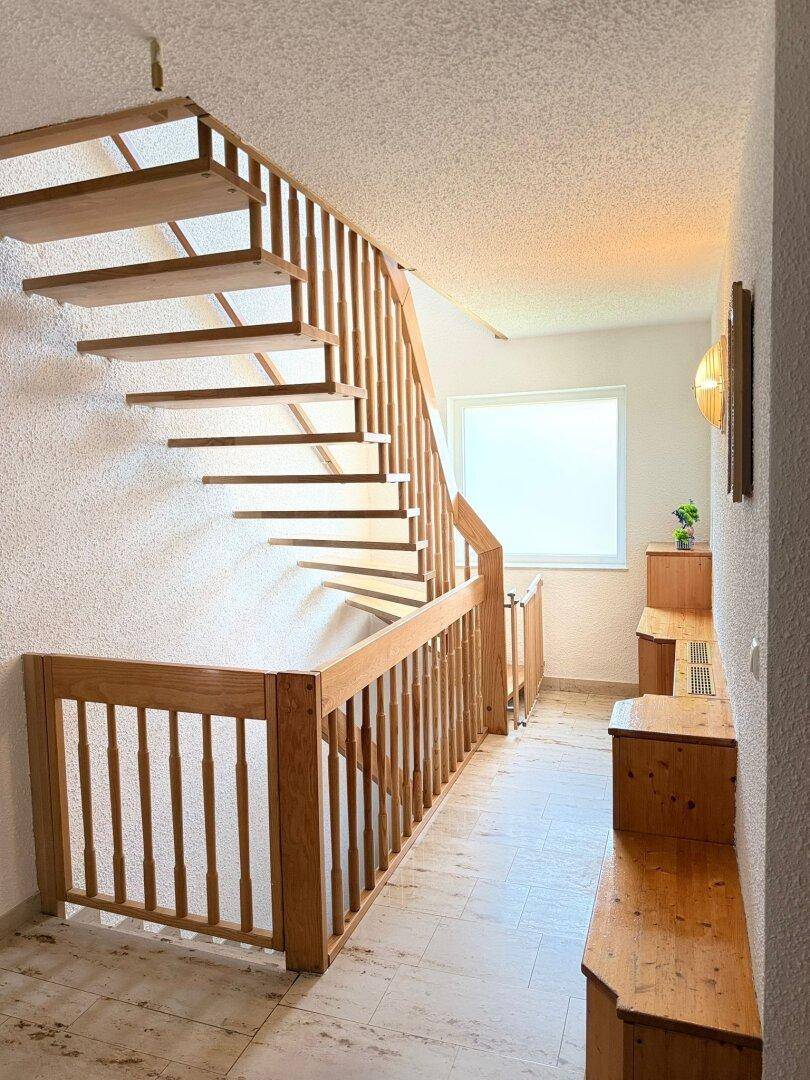 Heller Flur mit offener Holztreppe, Holzgeländer und hellen Fliesenböden.