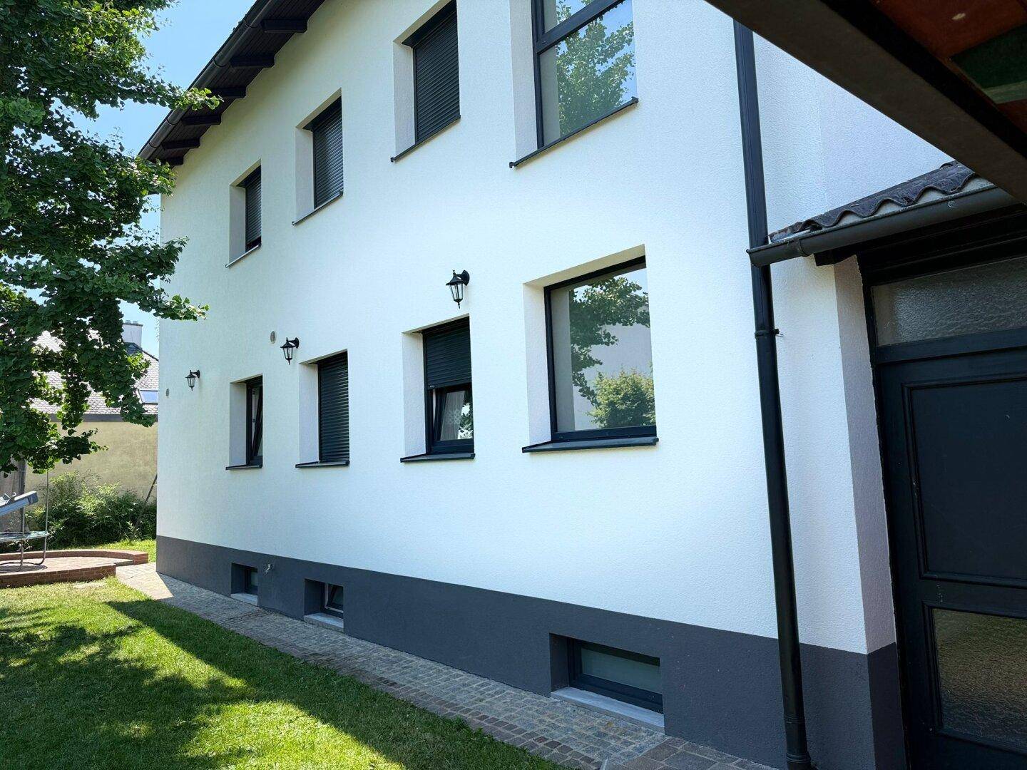 Seitenansicht des Hauses mit weißer Fassade, dunklen Fensterläden und angrenzendem Rasenbereich.