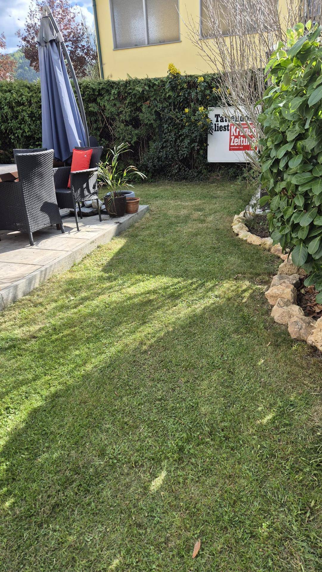 Sonniger Garten mit Rasenfläche, gepflasterter Terrasse und Sitzgelegenheiten unter einem Sonnenschirm.