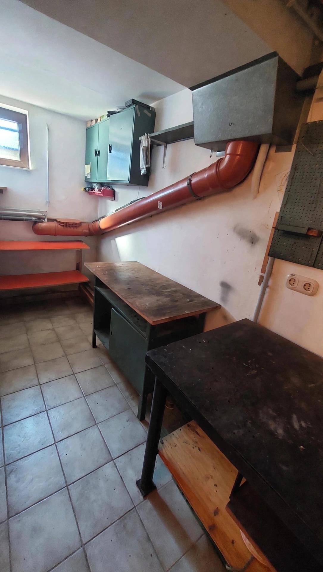 Werkstatt im Keller mit Arbeitstischen, Regalen und sichtbaren Rohren.