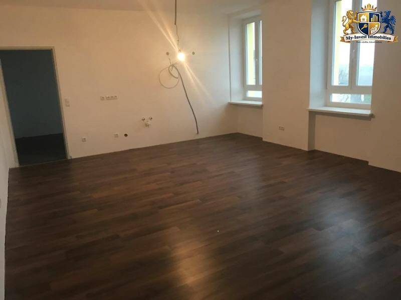 Geräumiger Wohnbereich mit dunklem Laminatboden und zwei hohen Fenstern, bereit für die Einrichtung.