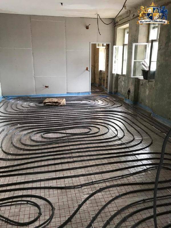 Innenraum im Bauzustand mit sichtbarer Fußbodenheizungsinstallation und Fenstern.
