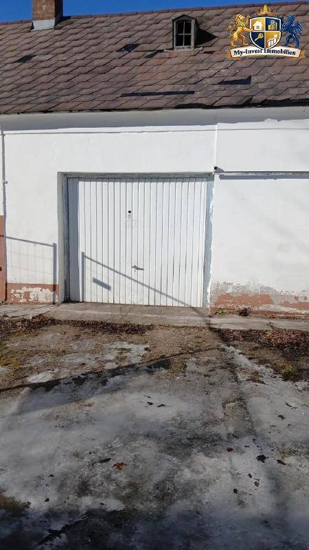 Außenansicht einer Garage mit weißem Tor und Zufahrt, Teil eines älteren Gebäudes mit Ziegeldach.