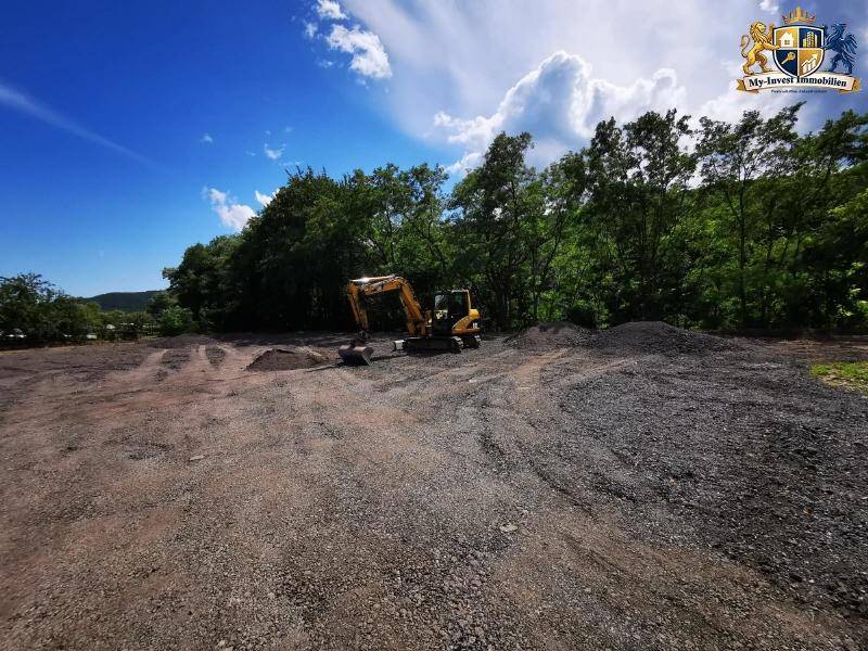 Bagger auf dem vorbereiteten Baugrundstück umgeben von grünen Bäumen unter strahlend blauem Himmel.