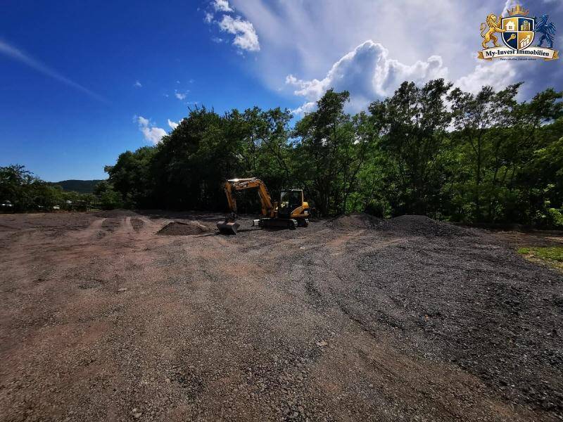 Bagger auf dem vorbereiteten Baugrundstück mit Erdhaufen und Wald im Hintergrund an einem sonnigen Tag.
