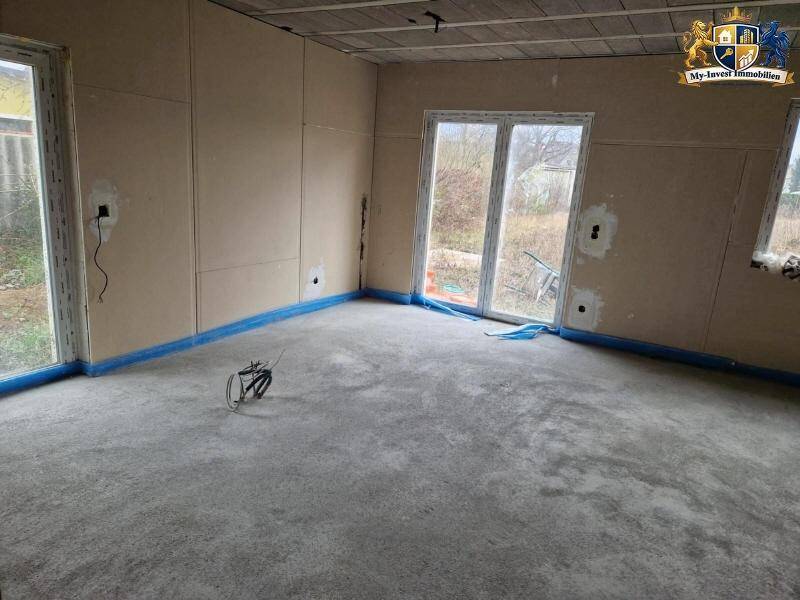 Rohbau eines Raumes mit Trockenbauwänden, Betonboden und großen Glastüren, die ins Freie führen.