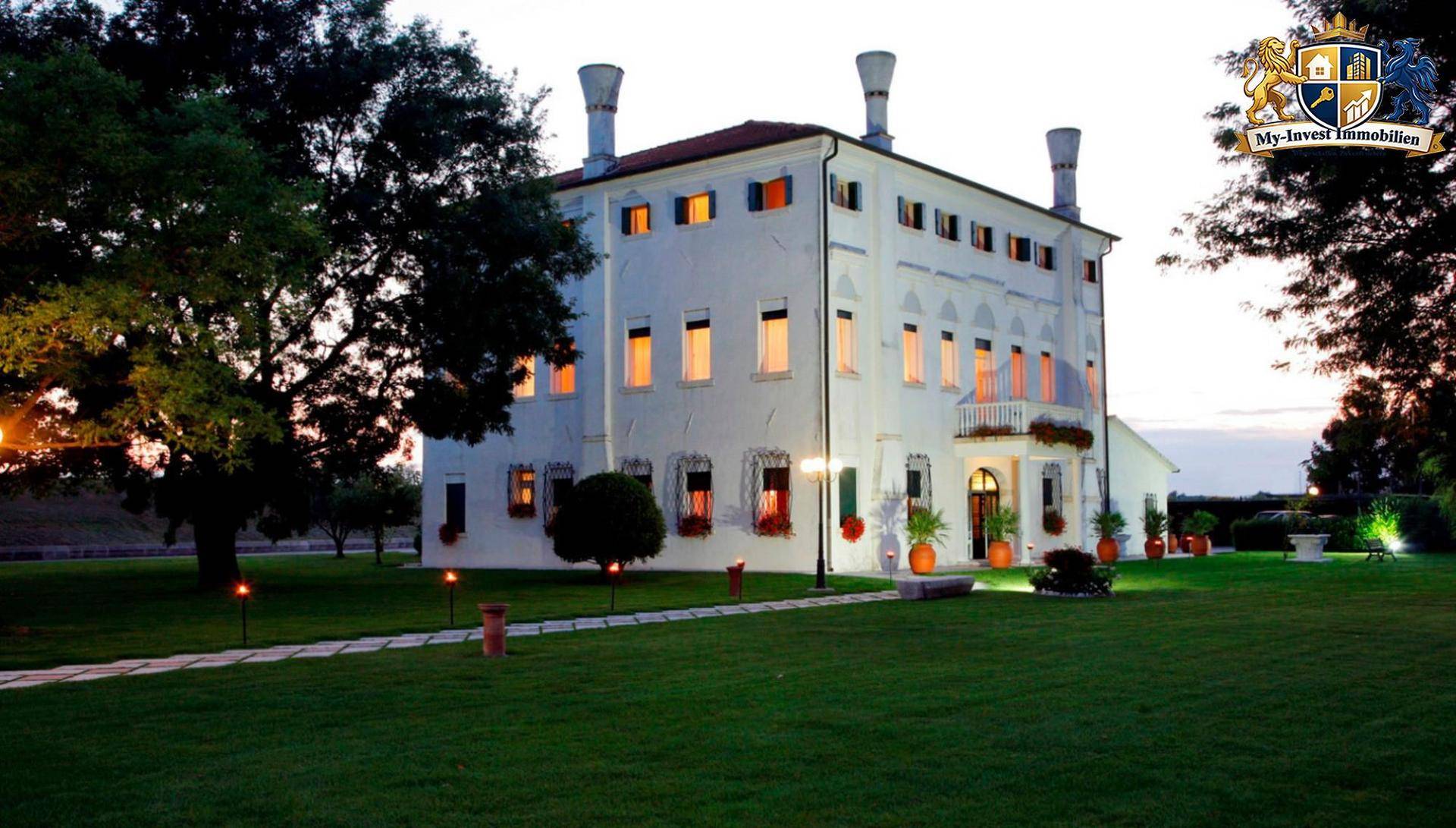 Historisches Gebäude in der Abenddämmerung mit beleuchteten Fenstern und einem gepflegten Vorgarten.