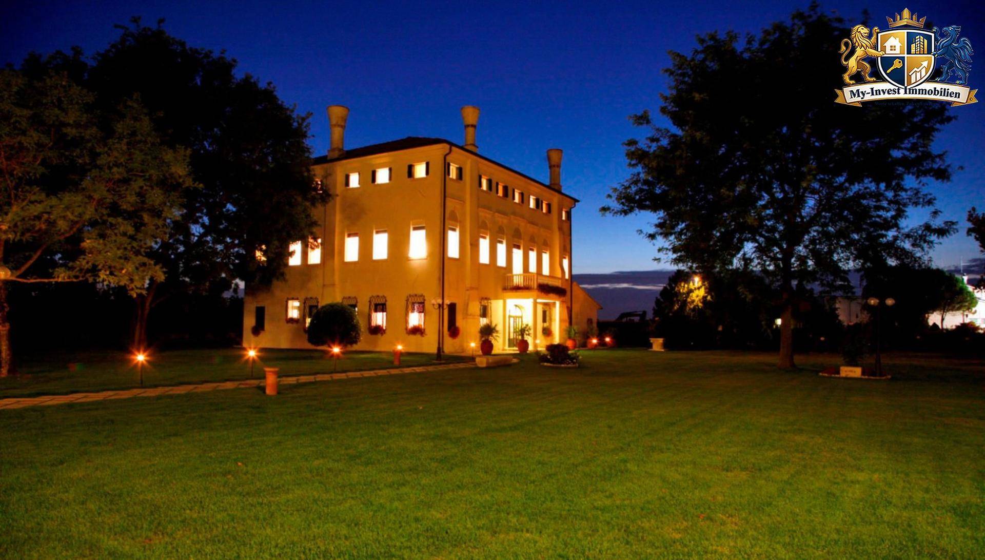 Beleuchtetes historisches Gebäude in der Nacht, umgeben von einem großen, dunklen Garten mit Wegbeleuchtung.