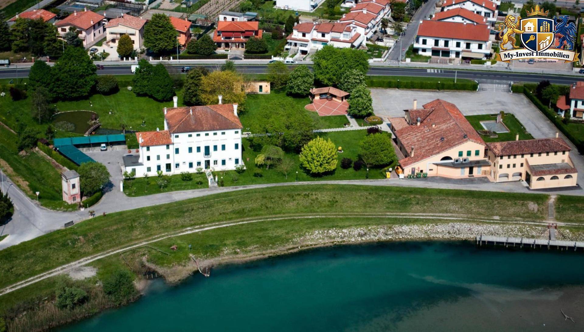 Luftaufnahme eines weitläufigen Anwesens mit Hauptgebäude, Nebengebäuden und einem Fluss oder Kanal.