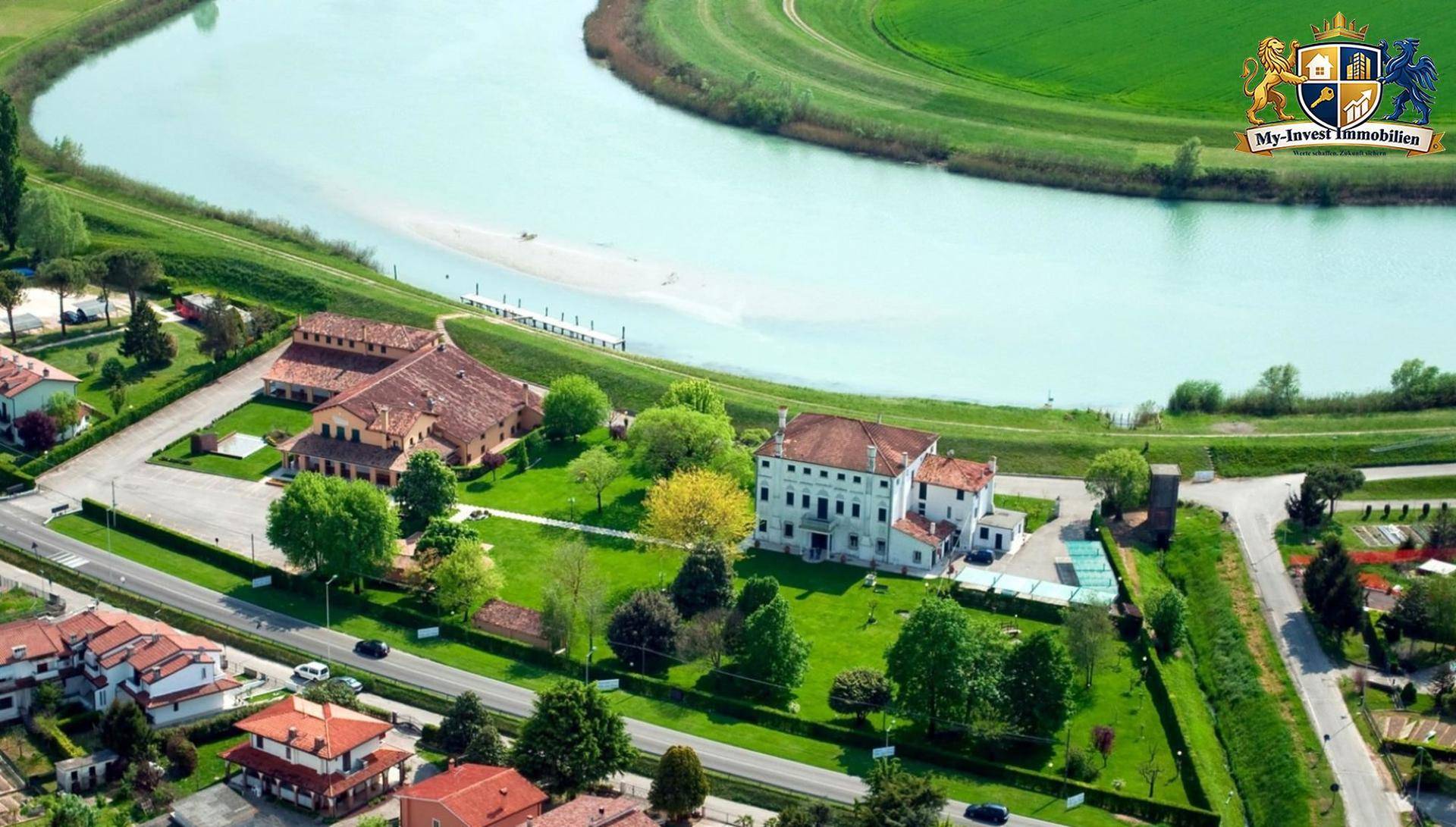 Weitläufiges Anwesen mit historischem Gebäude und Nebengebäuden, umgeben von Grünflächen und einem Fluss.