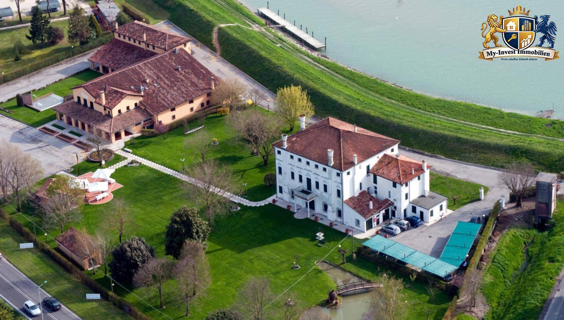Luftaufnahme eines weitläufigen Anwesens mit mehreren Gebäuden, großem Garten und angrenzendem Fluss oder See.